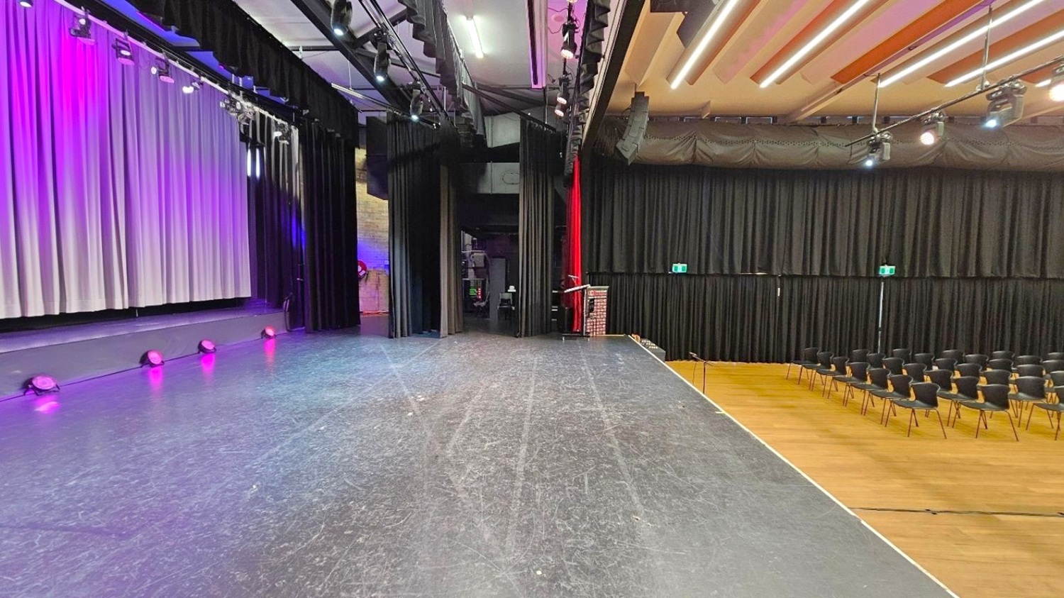 A stage with a black floor and overhead lighting. On the left, a large backdrop with text and purple lighting. In the centre, black curtains and a podium with a microphone. On the right, rows of black chairs on a wooden floor. The ceiling has exposed beams and lighting fixtures.