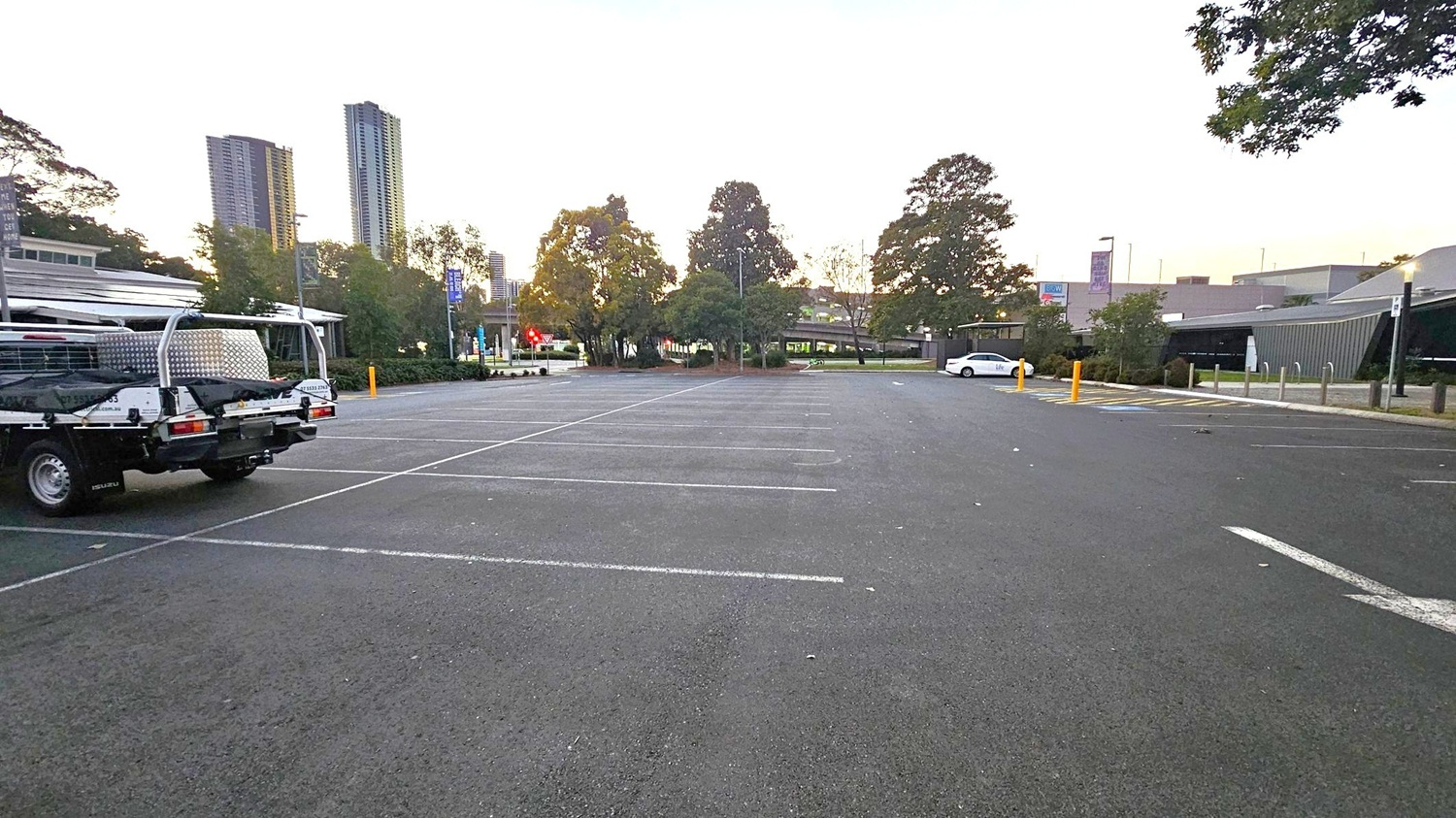 An empty car park with a few vehicles. On the left, a utility vehicle is parked, while on the right, a white car is visible. The background features tall buildings and trees, with a few signs and a low building on the right. The ground is asphalt with marked parking spaces, and there are a few yellow bollards near the white car. The sky is a gradient of light colours, suggesting early morning or late afternoon.