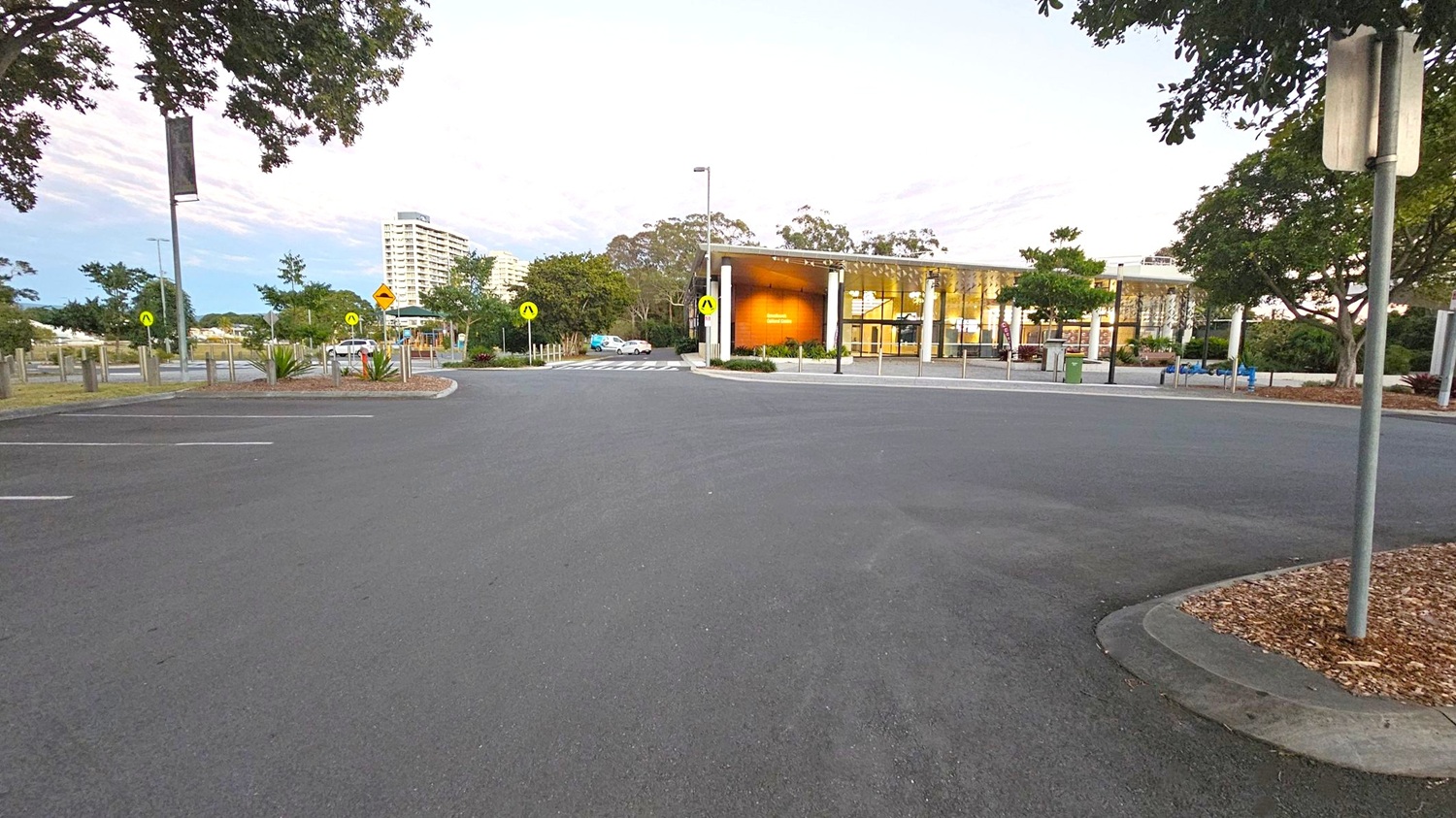 A wide, empty car park with a smooth asphalt surface. In the background, a modern building with large glass windows and a wooden facade is visible. To the left, there are trees and a few parked cars. On the right, more trees and a signpost are present. The area is well-lit with street lamps, and there are pedestrian crossing signs near the building. A tall building is visible in the distance.