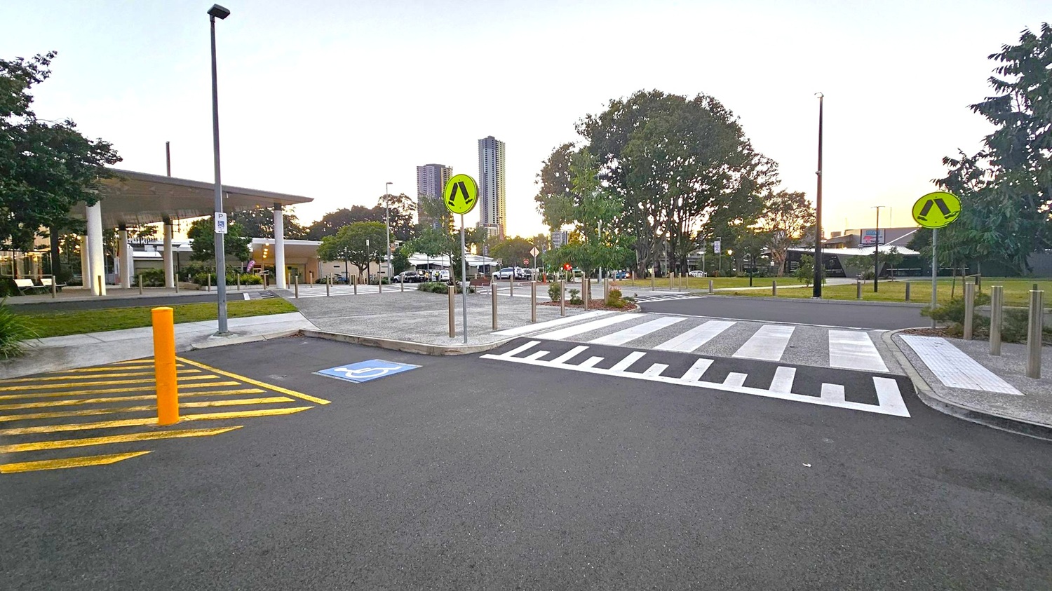 A paved area with a zebra crossing in the centre, flanked by pedestrian signs on poles. On the left, a building with a modern design and a sloped roof is visible, surrounded by trees. A yellow-striped area and a blue disabled parking sign are on the asphalt. In the background, tall buildings rise above the treeline, and the sky shows a hint of sunset. The foreground features a smooth, dark road surface.