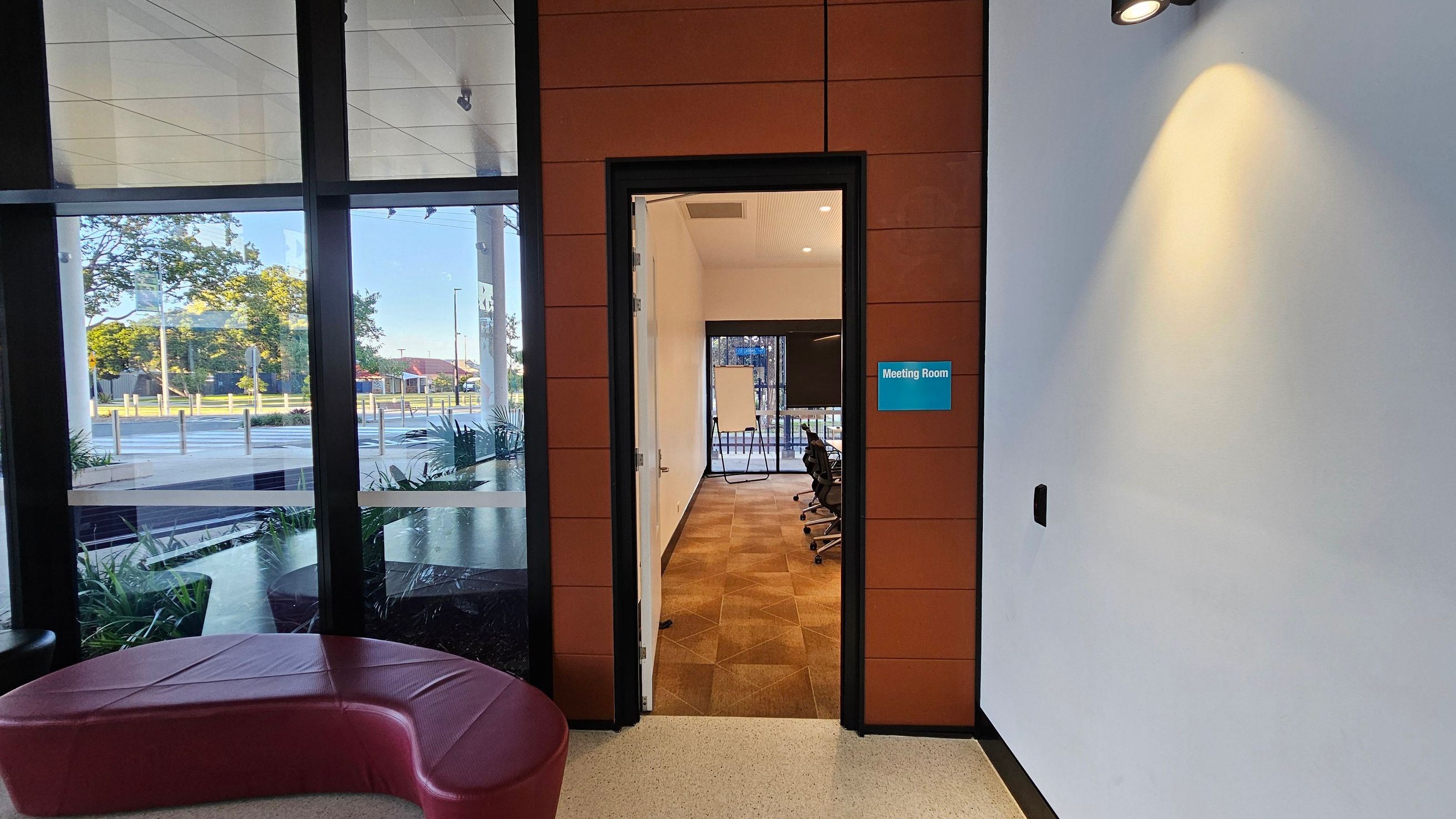 A hallway with a view into a meeting room through an open door. The meeting room has a brown tiled floor and several office chairs around a table. A whiteboard is visible at the far end. To the left, large windows show an outdoor scene with trees and a street. A curved red bench is positioned near the windows. The wall on the right is white with a spotlight casting light on it, and a sign reading "Meeting Room" is mounted beside the door.