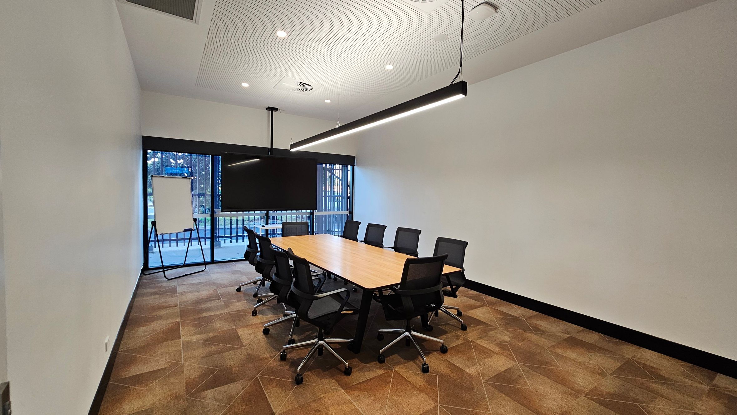 A modern conference room with a long wooden table surrounded by black office chairs on wheels. On the left, a flip chart stands near a large window with vertical blinds, offering a view of trees outside. A large flat-screen TV is mounted on the wall behind the table. The ceiling is white with recessed lighting and a long, black pendant light fixture. The floor is covered with geometric-patterned carpet tiles in shades of brown.
