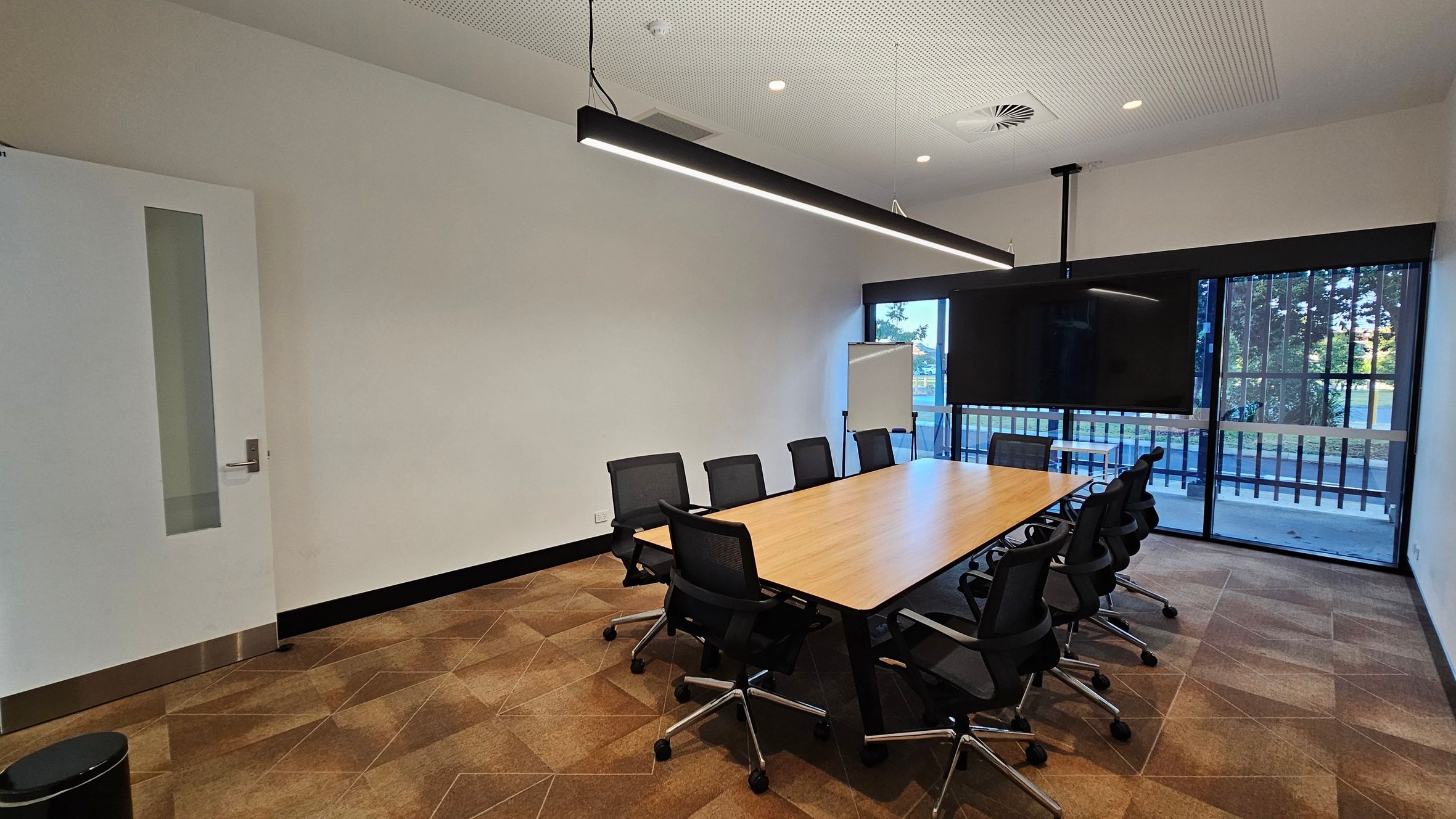 A modern conference room with a long wooden table surrounded by black office chairs on a patterned carpet floor. On the right, large windows with vertical bars offer a view of trees outside. A large flat-screen TV is mounted on the wall. A flip chart is positioned near the windows. The ceiling is white with recessed lighting and a linear pendant light hangs above the table. A door with a vertical window is on the left, and a small waste bin is nearby.