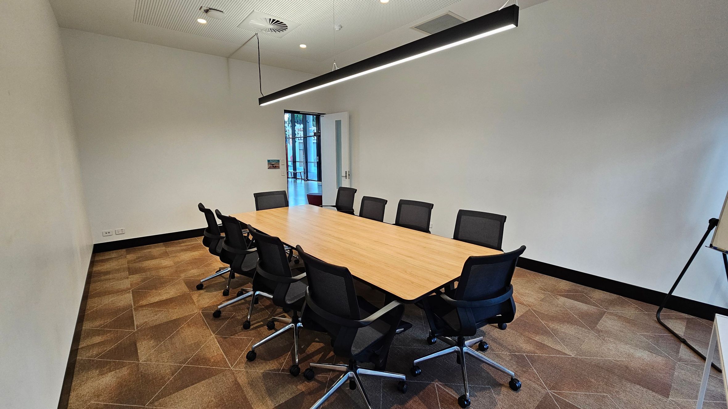 A conference room with a long wooden table in the centre, surrounded by ten black office chairs on wheels. The floor is covered with a patterned carpet in shades of brown. On the right, a whiteboard on a stand is partially visible. The ceiling features recessed lighting and a long, black hanging light fixture above the table. At the back, a door is open, leading to a hallway with glass panels. The walls are plain and light-coloured.