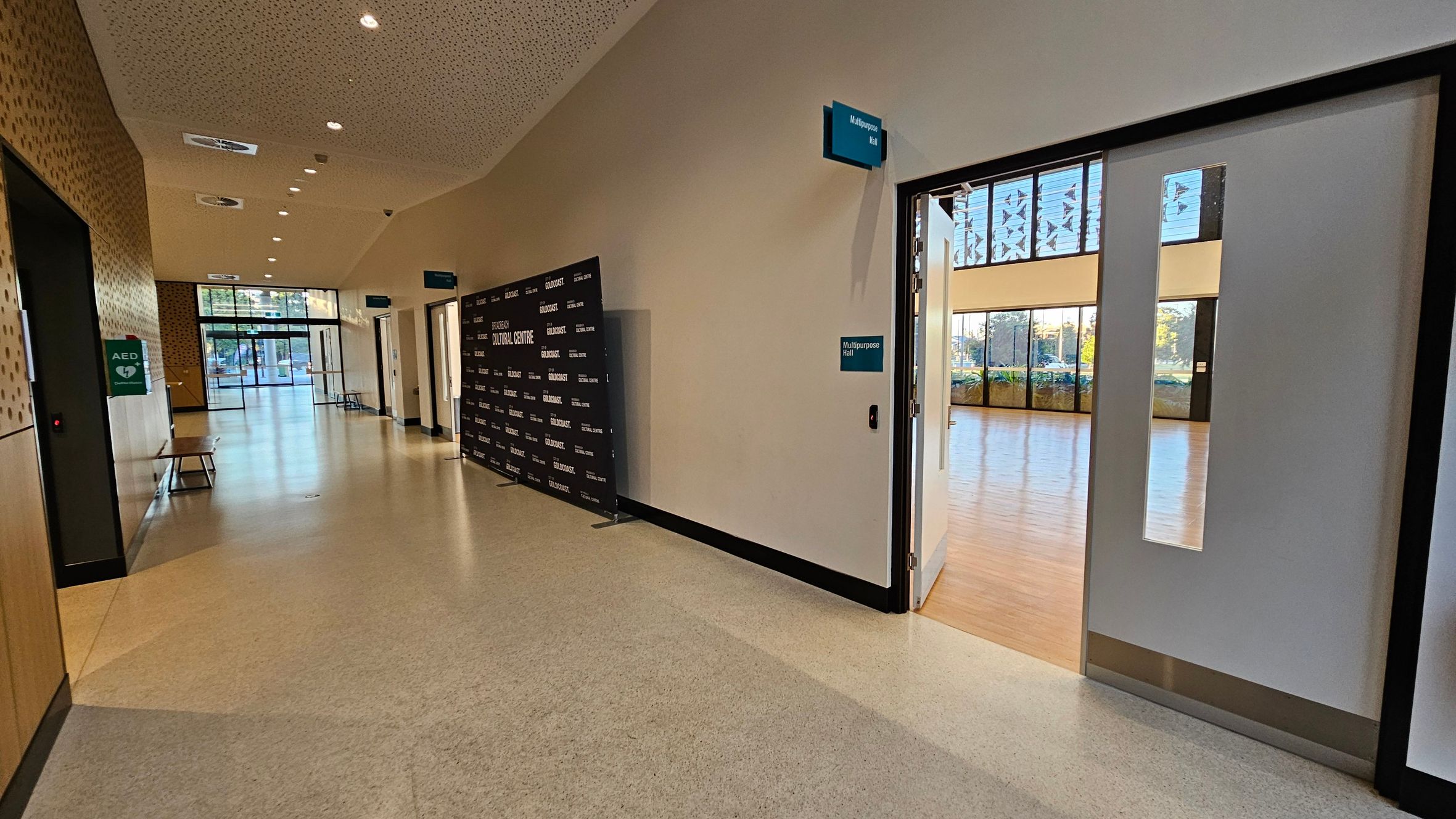 A hallway with a light-coloured floor and a perforated ceiling with recessed lights. On the left, there is a wall with a green AED sign and a bench. At the end of the hallway, glass doors lead outside. On the right, a door is open to a multipurpose hall with wooden flooring and large windows. A black backdrop with white text is positioned against the right wall. Signs for the multipurpose hall are visible above and beside the door.