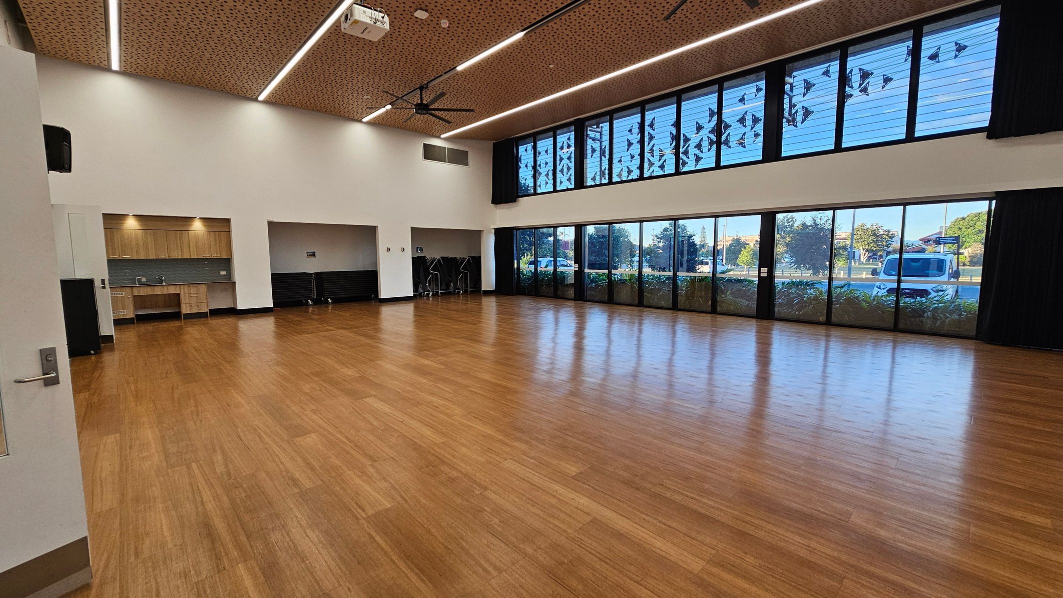 A spacious room with a wooden floor and a perforated wooden ceiling featuring linear lights and a ceiling fan. On the left, there is a kitchenette with wooden cabinets and a sink. Adjacent to it is a storage area with stacked chairs. The right side has large windows and glass doors, allowing natural light and views of trees and a parked van outside. The walls are white, and the room appears to be a multi-purpose space.