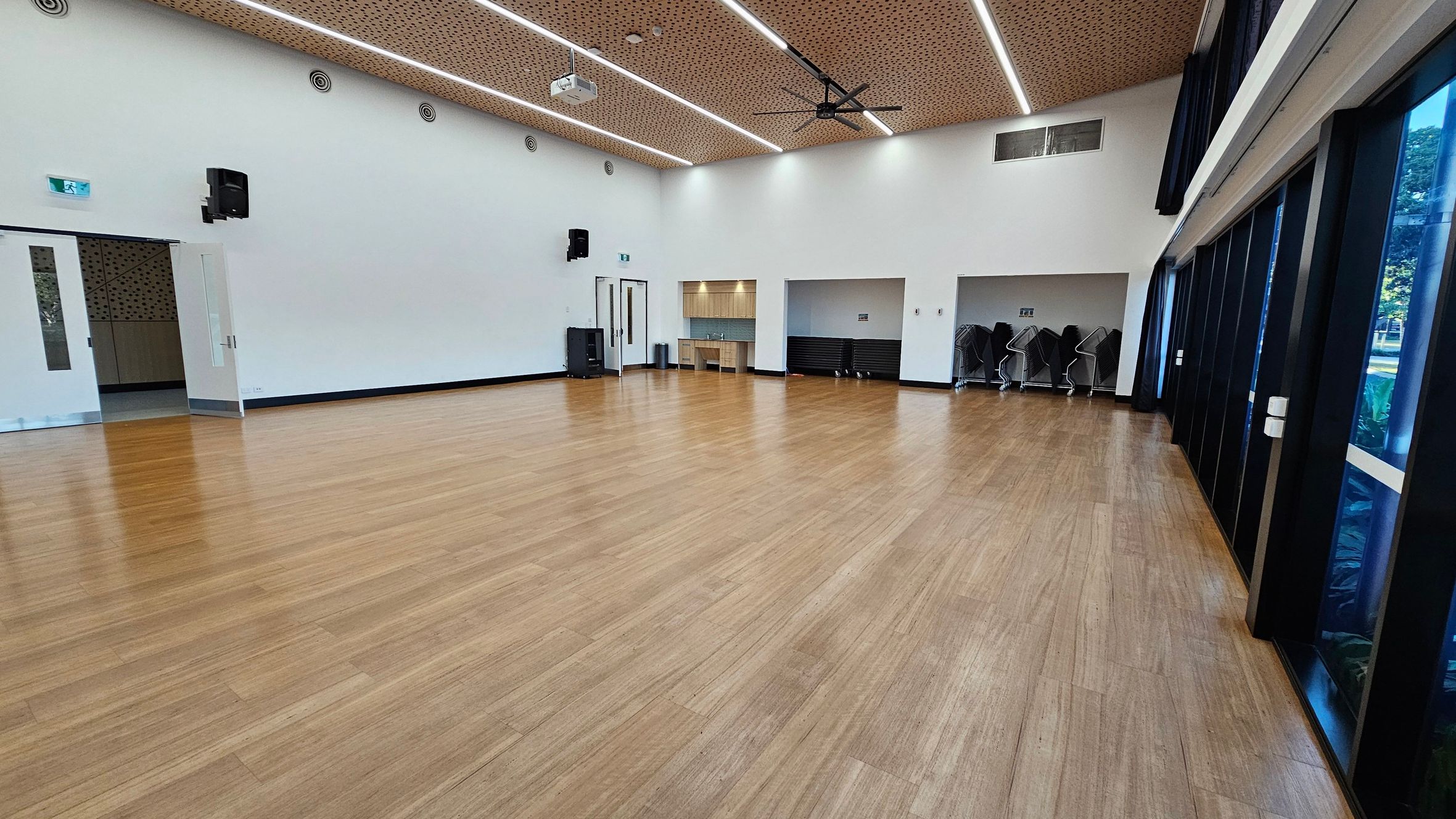 A spacious room with a wooden floor and a perforated wooden ceiling featuring linear lights and a large ceiling fan. On the left, there are double doors and a speaker mounted on the wall. In the centre, a small kitchenette with a sink is visible, and to its right, a storage area with stacked chairs. The right side has large windows with dark frames, allowing natural light to enter.