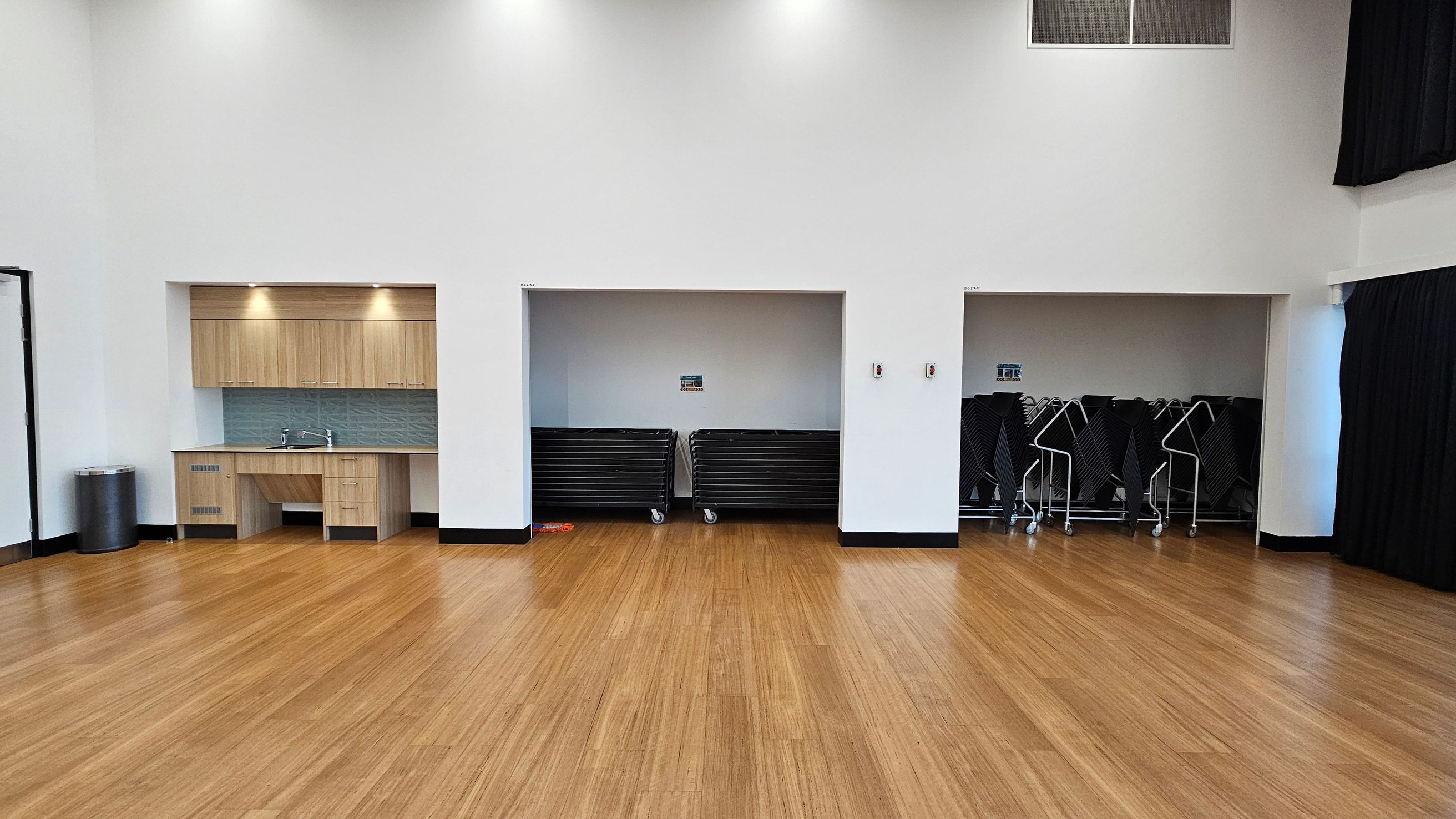 A spacious room with a wooden floor and white walls. On the left, there is a kitchenette with light wood cabinets, a sink, and a backsplash. Next to it is a metal bin. In the centre, there are two storage alcoves; the left one contains stacked tables on wheels, and the right one has folded chairs on trolleys. The ceiling is high with recessed lighting, and there are black curtains partially visible on the right side.