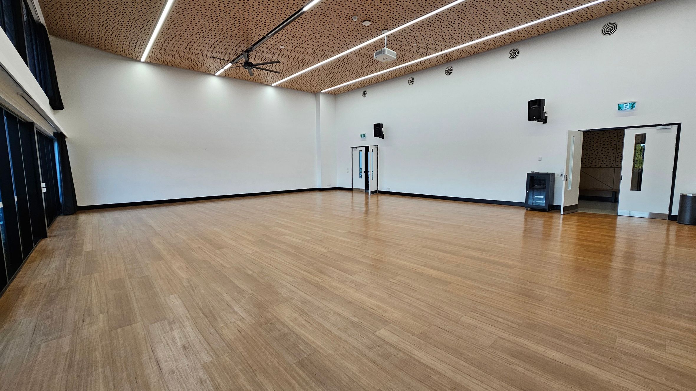 A spacious room with a light wooden floor and white walls. On the left, there are large windows with dark curtains. The ceiling is wooden with linear lights and a large fan. On the right, there are two doors, one open and one closed, with a small black speaker mounted on the wall above. A waste bin and a small cabinet are also on the right side. The room has a modern and minimalistic design.