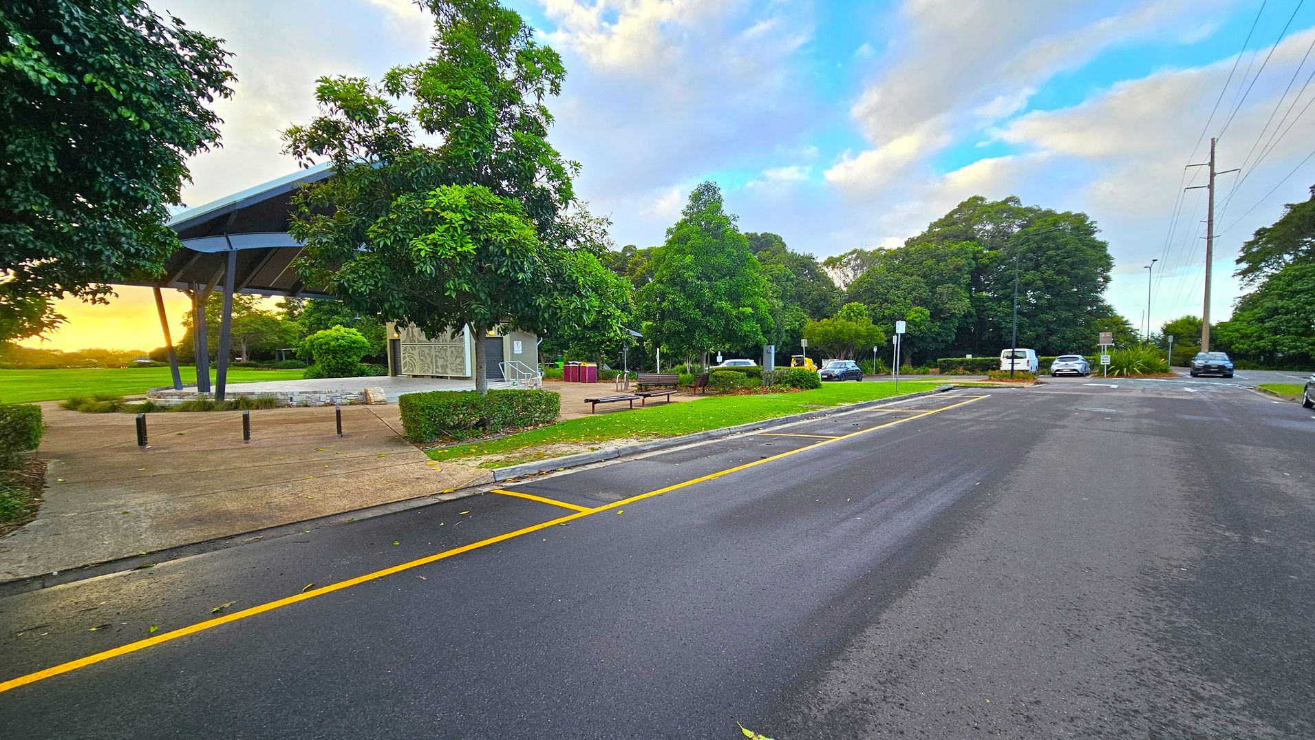 A paved road runs through the right side, with yellow lines marking parking spaces. On the left, there's a covered structure with a slanted roof, surrounded by trees and greenery. Benches and a small path are visible near the structure. Several cars are parked along the road, and utility poles with wires run parallel to the street. The background features more trees and a grassy area, with a hint of sunset light visible.