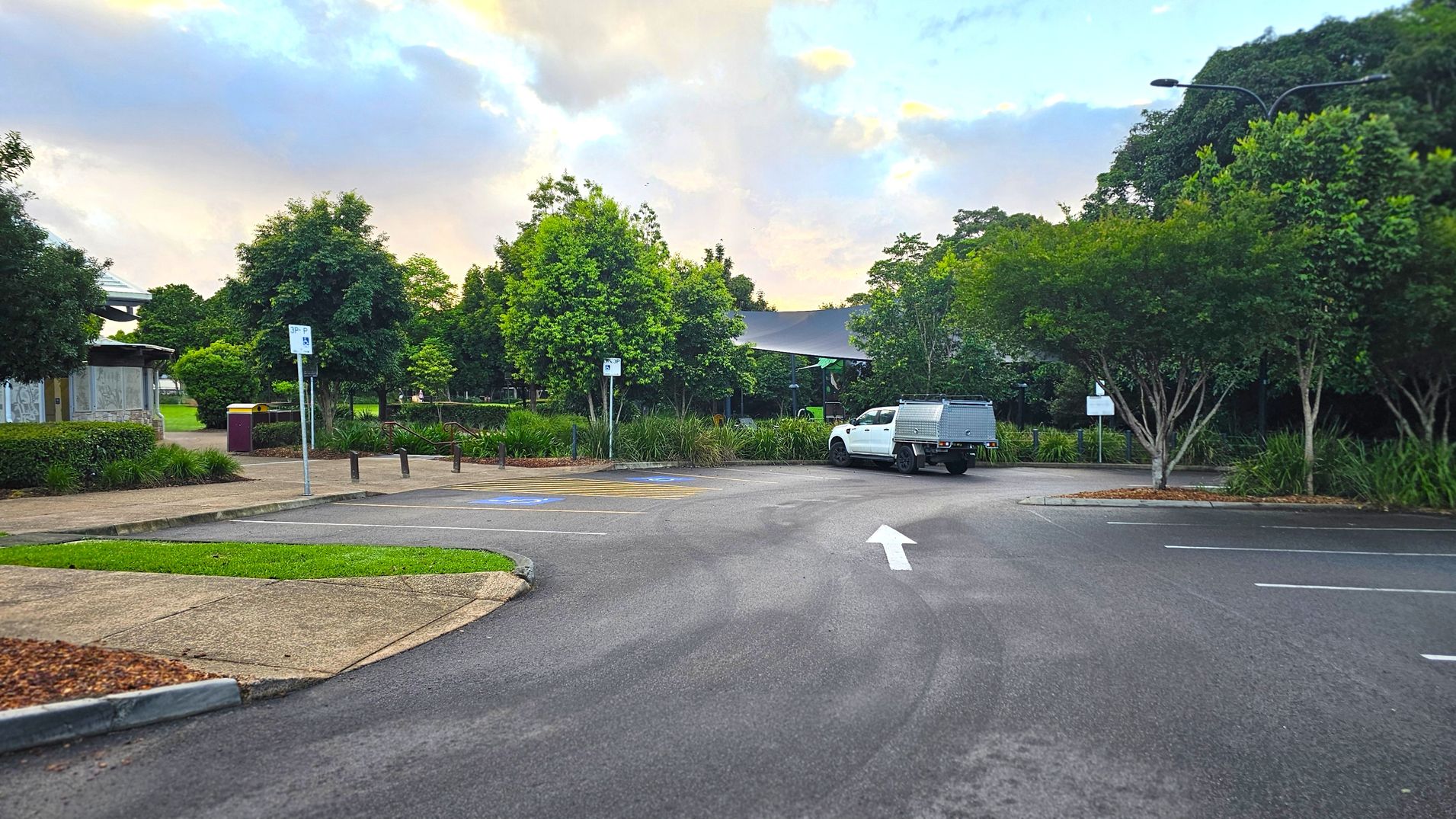 A car park with a white utility vehicle parked on the right. The ground is paved with marked parking spaces and a directional arrow pointing forward. On the left, there's a footpath bordered by grass and a small building partially visible. Trees and shrubs line the background, providing greenery. A signpost is visible near the centre, and a streetlamp is on the right.