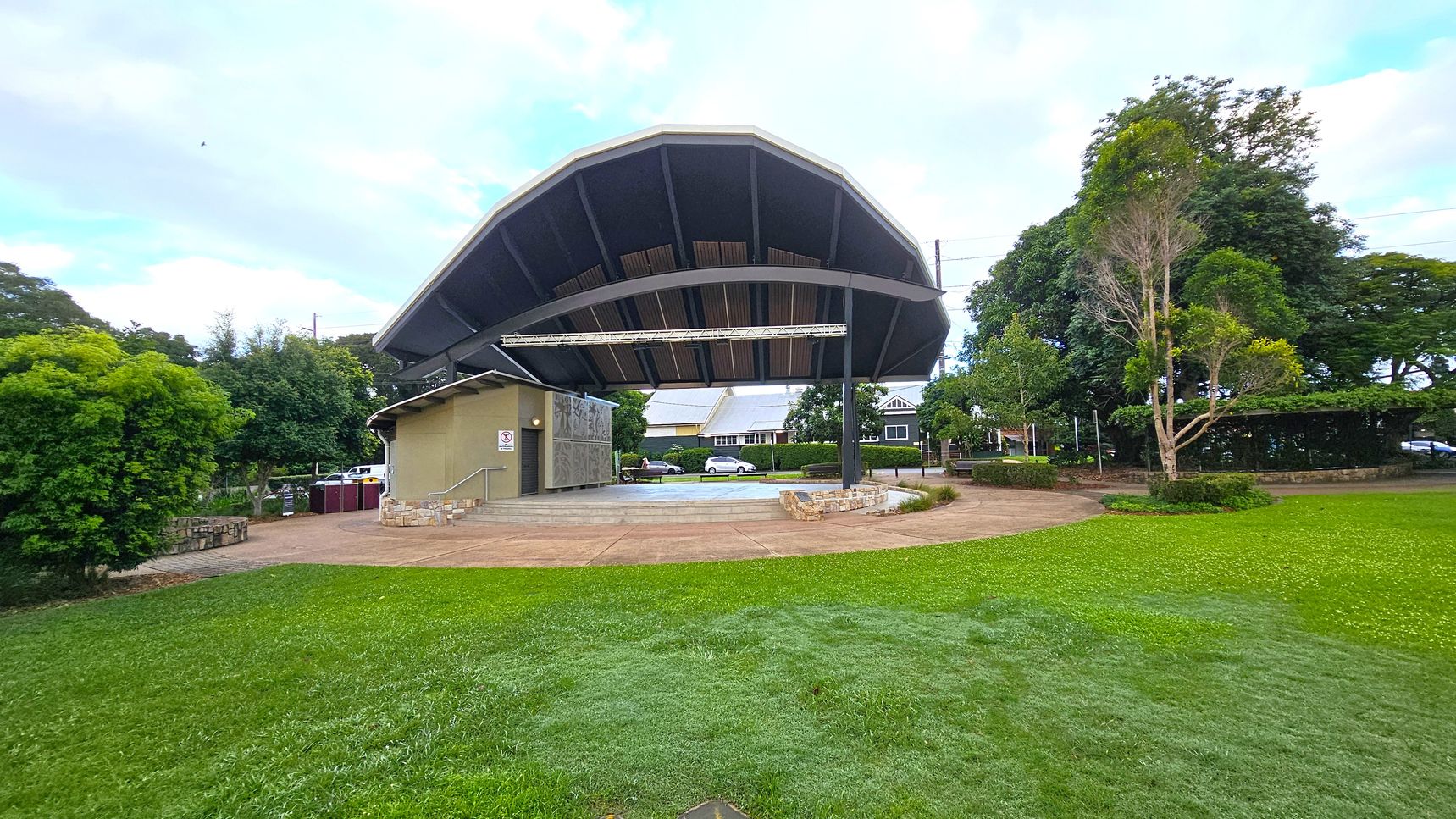 A large, open-air stage with a curved roof is in the centre, surrounded by a grassy area. The stage has a beige building attached to it on the left, with steps leading up. Trees and shrubs are on both sides, with a few small trees on the right. In the background, there are residential houses and a parked car. The ground is a mix of grass and paved pathways.