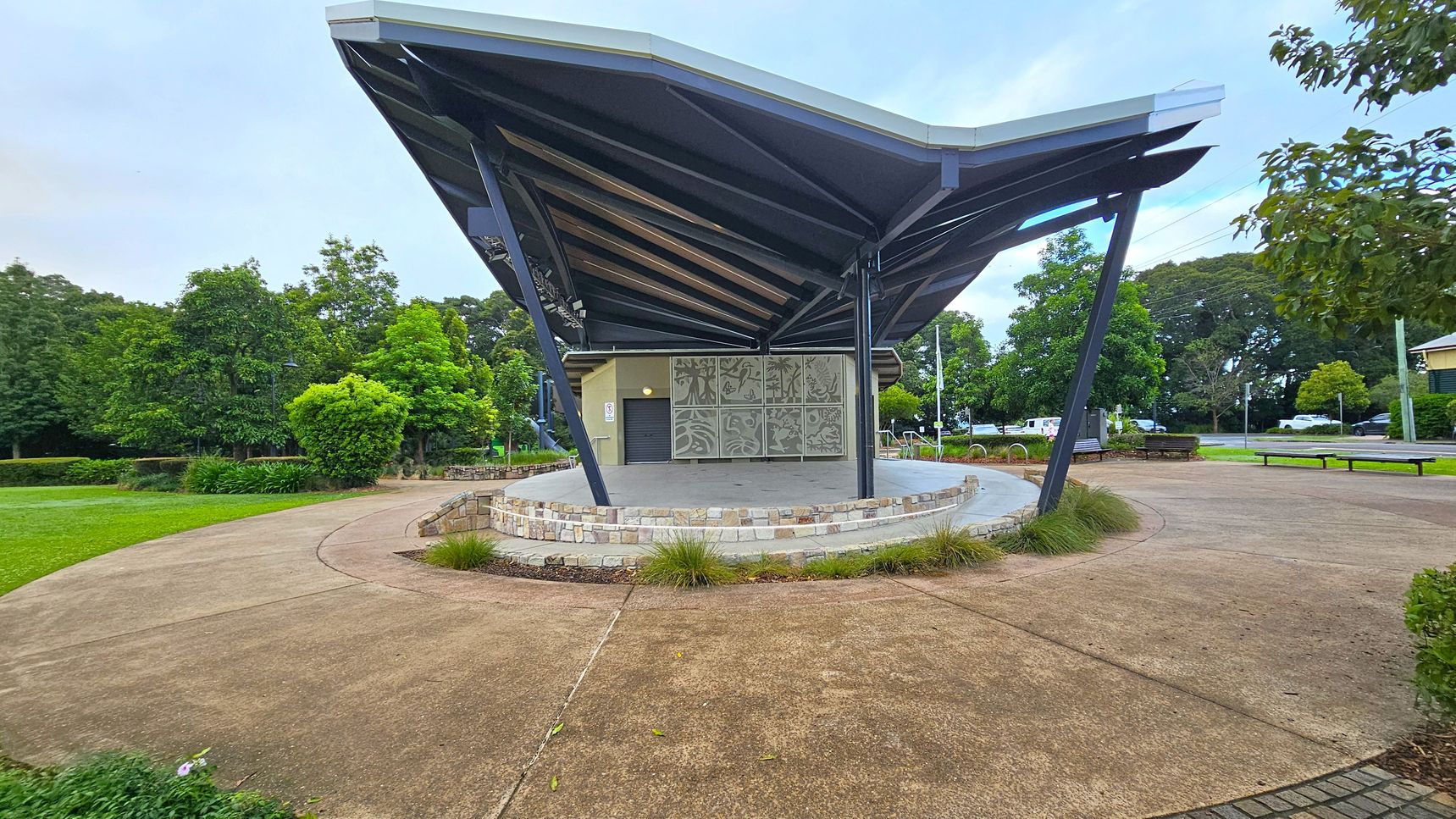 A modern outdoor stage with a distinctive angular roof supported by dark metal beams. The stage has a stone base and decorative panels at the back. Surrounding the stage is a paved area with patches of grass and small shrubs. To the left, there are lush green trees and a grassy lawn. On the right, a few benches and more trees are visible, along with a road in the background. The sky is overcast, creating a soft light over the scene.