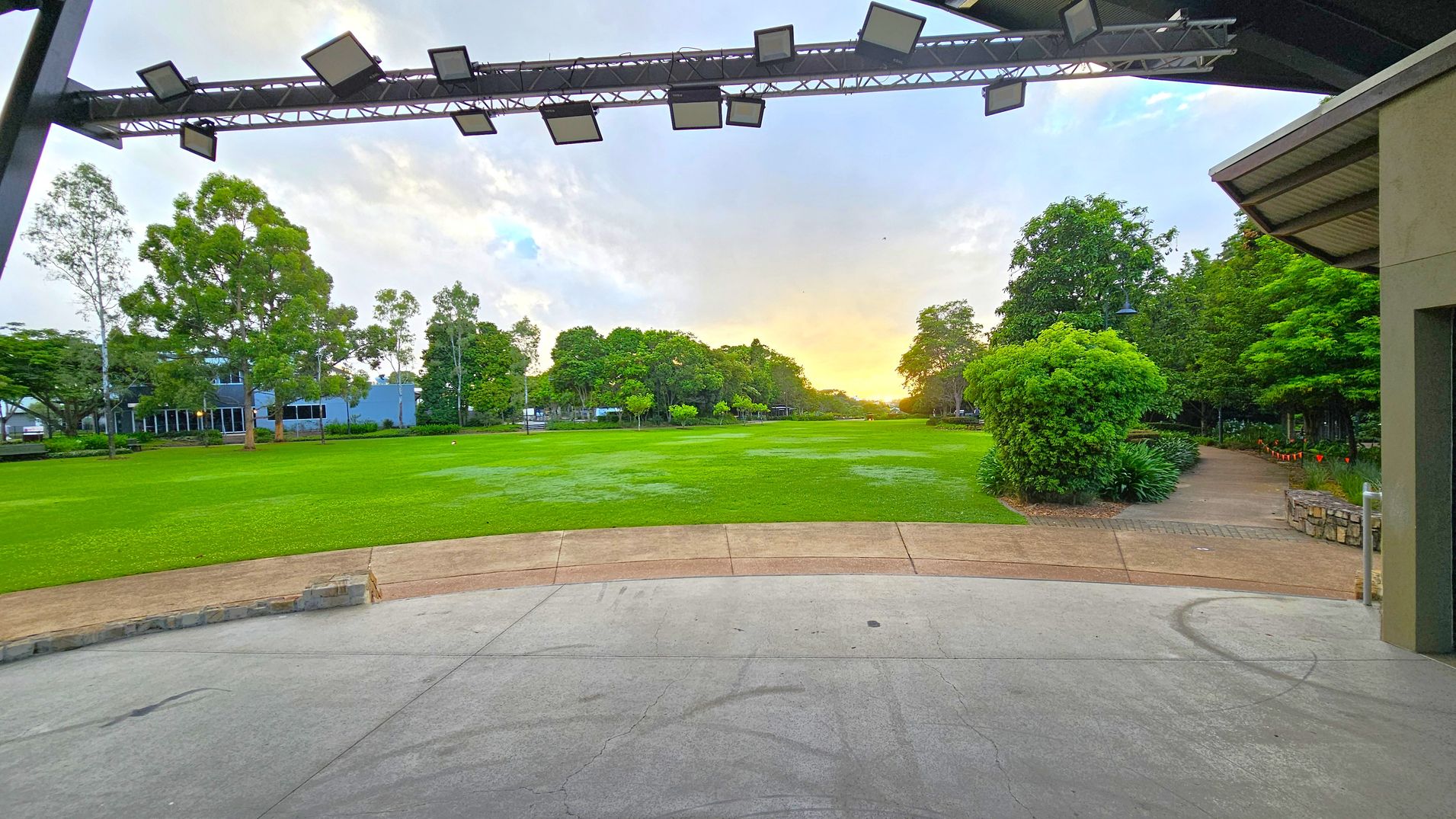 A wide, open grassy field stretches out in the centre, bordered by lush green trees. On the left, there are buildings partially visible behind the trees. On the right, a pathway lined with bushes and a stone wall leads into the distance. Overhead, a metal structure with lights is visible. The foreground features a concrete surface with a curved edge leading to the grass. The sky is bright near the horizon, suggesting early morning or late afternoon light.