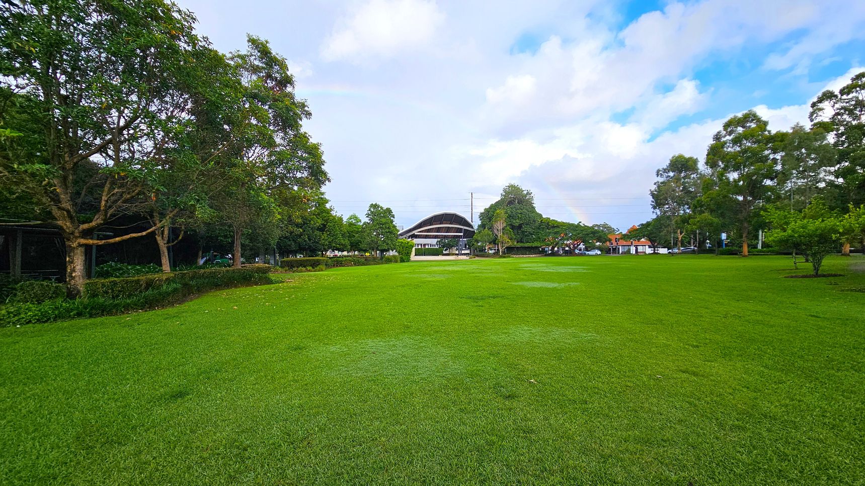 A large, open grassy field stretches across the foreground. On the left, there are several trees with dense foliage, and a hedge runs parallel to them. In the centre background, a modern, curved-roof structure is visible, surrounded by more trees. To the right, additional trees and a few parked cars can be seen. The scene is well-lit, suggesting daytime.