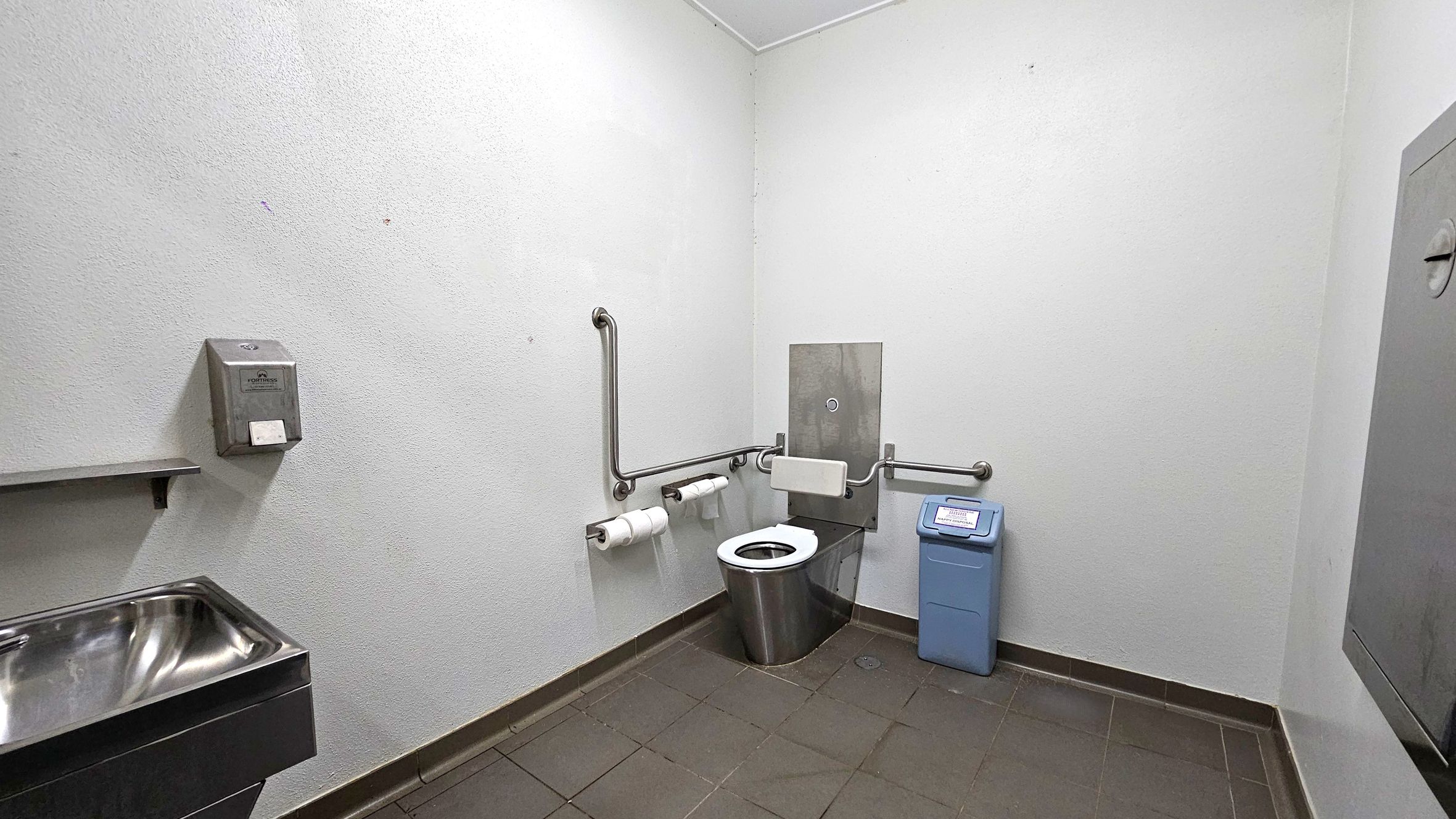 A public restroom with a stainless steel toilet in the centre, equipped with grab bars on both sides. To the right, a blue sanitary bin is placed on the tiled floor. On the left, a stainless steel sink is mounted on the wall, with a soap dispenser above it. The walls are plain and light-coloured, and the floor is covered with dark tiles. The ceiling is plain and light-coloured.