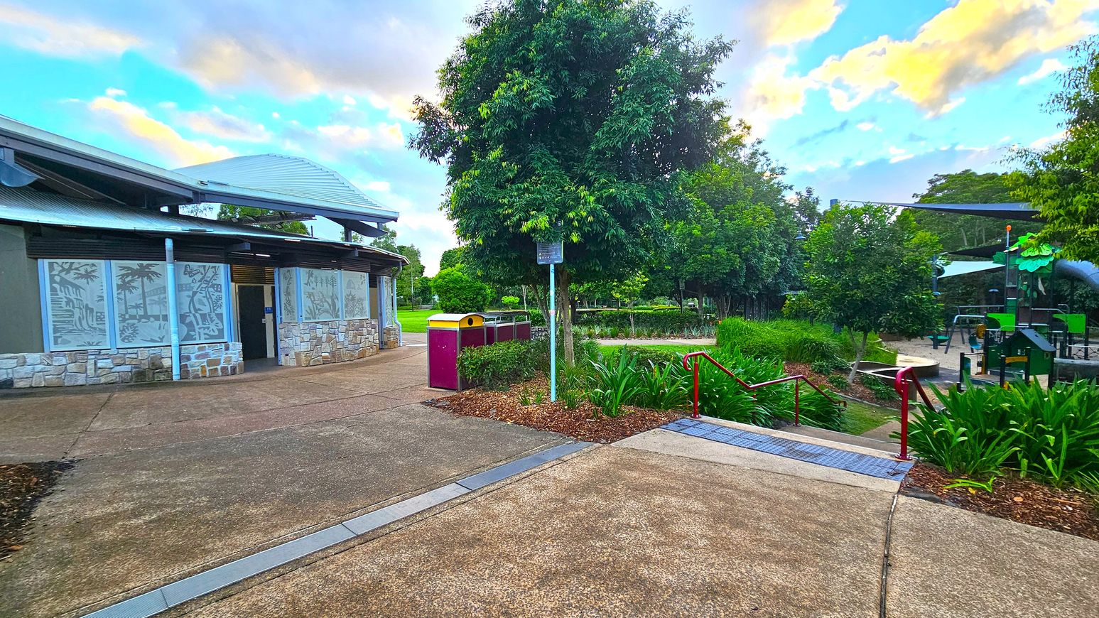 A paved pathway leads through a park area. On the left, there's a building with a stone base and decorative panels. In the centre, a large tree stands next to a red and yellow bin. On the right, a playground with green equipment is partially visible, surrounded by lush greenery. Red railings border a small set of steps leading downwards. The area is well-maintained with various plants and trees.