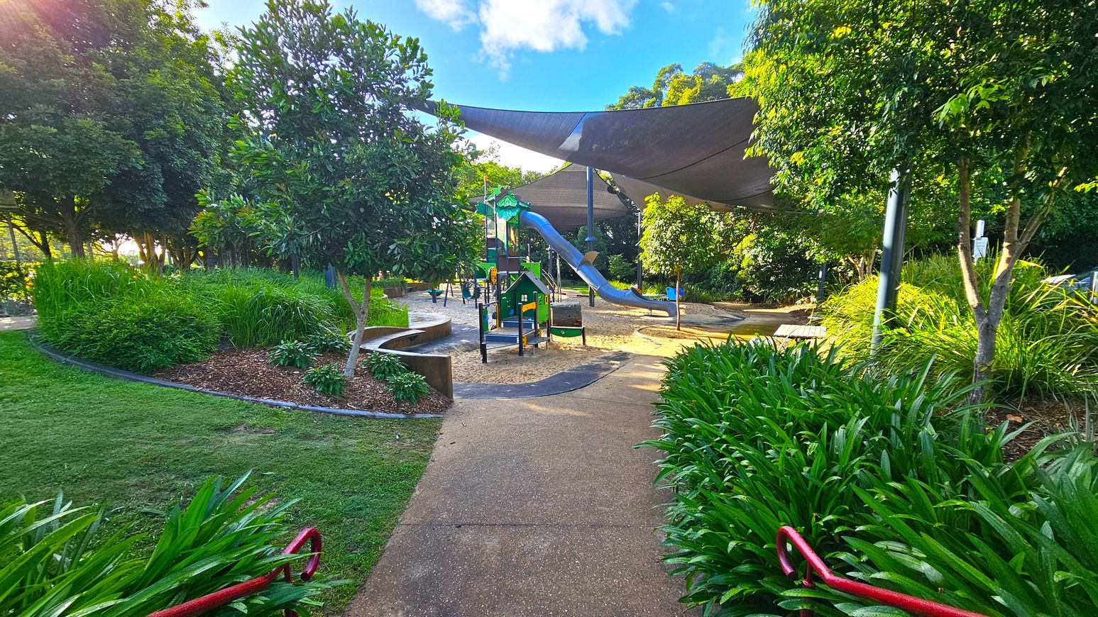 A playground surrounded by lush greenery. In the centre, there is a play structure with slides and climbing elements, set on a sandy area. Overhead, large shade sails provide cover. On the left, dense bushes and trees create a natural border, while on the right, more trees and a path lined with tall grass are visible. The foreground features a paved path with red railings, leading into the play area.