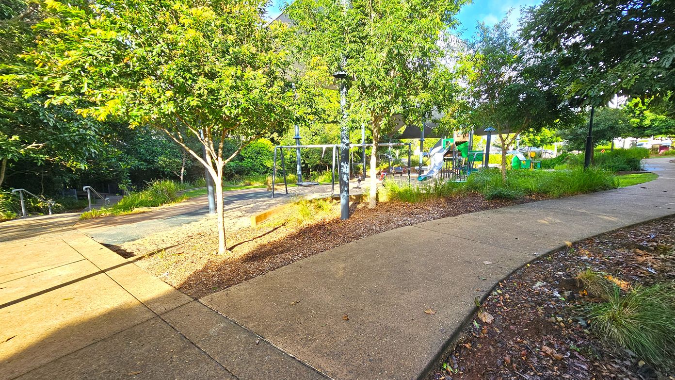 A park scene with a paved pathway curving from the foreground to the right. On the left, there are trees with green foliage and a set of steps with metal railings. In the centre, a playground area features swings and a slide, surrounded by more trees and grass. The ground is covered with mulch and grass, and there are lamp posts scattered throughout the area. The background is filled with dense greenery.