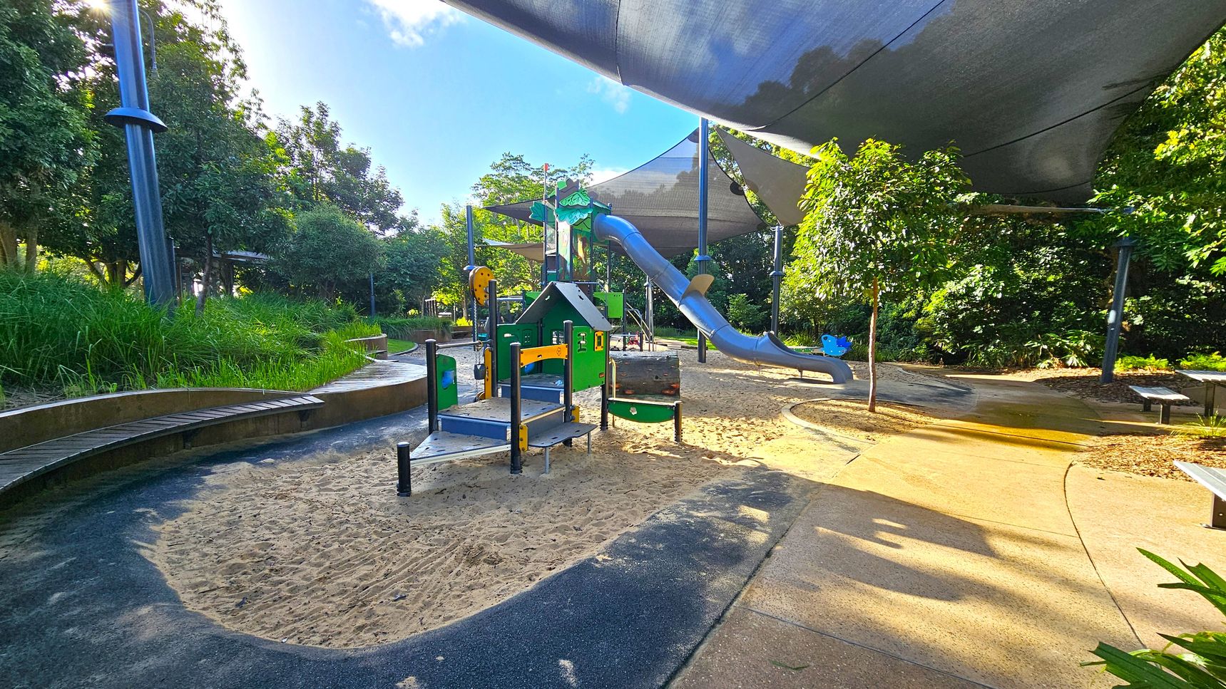 A playground with a sandy area in the foreground, featuring a small climbing structure with steps and a slide. To the right, a larger blue slide extends from a higher platform. The ground is a mix of sand and paved paths. Overhead, large shade sails provide cover. Lush greenery and trees surround the area, with a bench visible on the right. The sunlight filters through the trees, casting shadows on the ground.