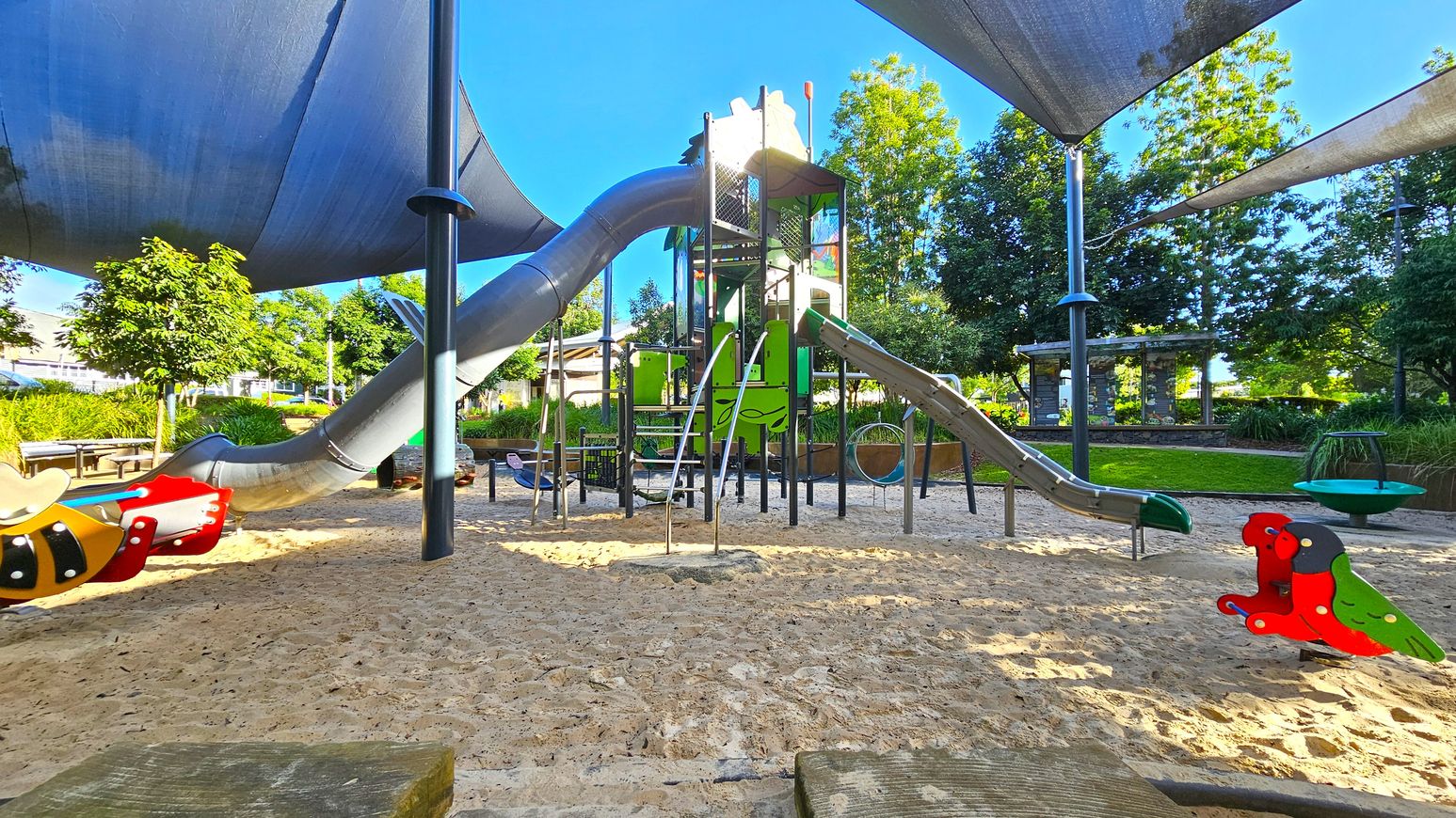 A playground with a sandy floor features various play equipment. In the centre, there is a tall climbing structure with slides, including a large tube slide. To the left, a colourful spring rider shaped like a bee is visible. On the right, another spring rider resembles a parrot. The area is shaded by large fabric canopies supported by poles. In the background, there are trees and a grassy area with benches.