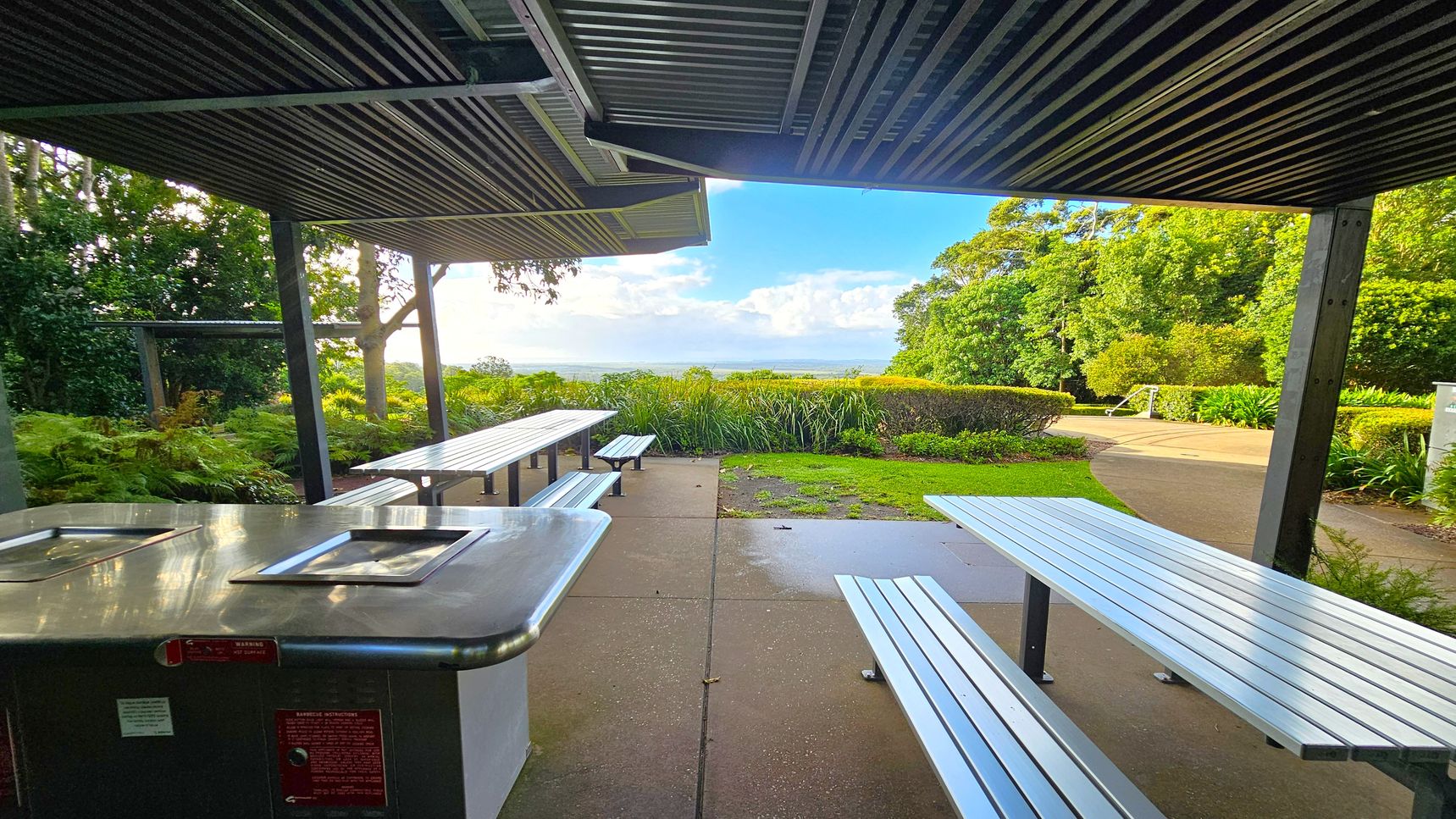 A covered outdoor area with metal picnic tables and benches on a concrete floor. On the left, there's a stainless steel barbecue unit. The ceiling is made of corrugated metal. In the background, lush greenery and trees surround the area, with a view of the horizon and a partly cloudy sky. A pathway curves through the garden, bordered by bushes and grass.