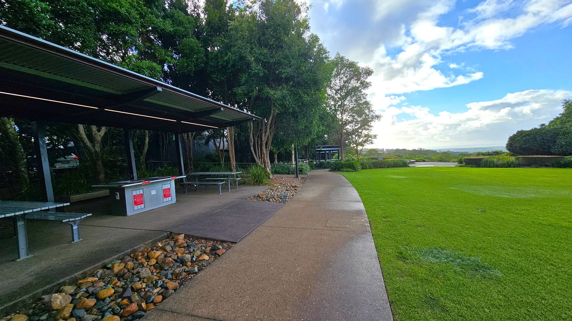 A park scene with a covered picnic area on the left, featuring metal benches and a barbecue station. The ground is paved with a mix of concrete and pebbles. Trees provide shade in the background. On the right, there's a well-maintained grassy area leading to a distant view of the horizon. The sky is partly cloudy, and the overall setting is open and spacious.