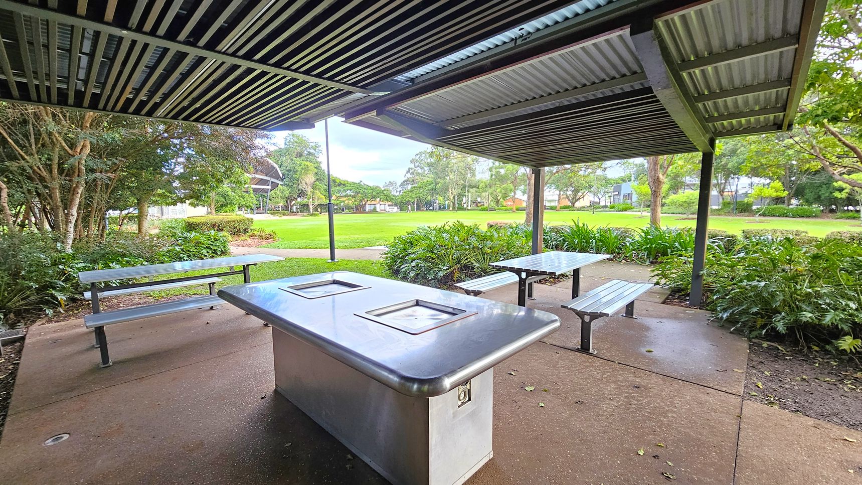 A covered outdoor area with a metal roof and a concrete floor. In the foreground, there is a stainless steel barbecue grill. To the left and right, there are metal picnic tables with benches. The background features a grassy park with trees and shrubs, and a path leading through the greenery.
