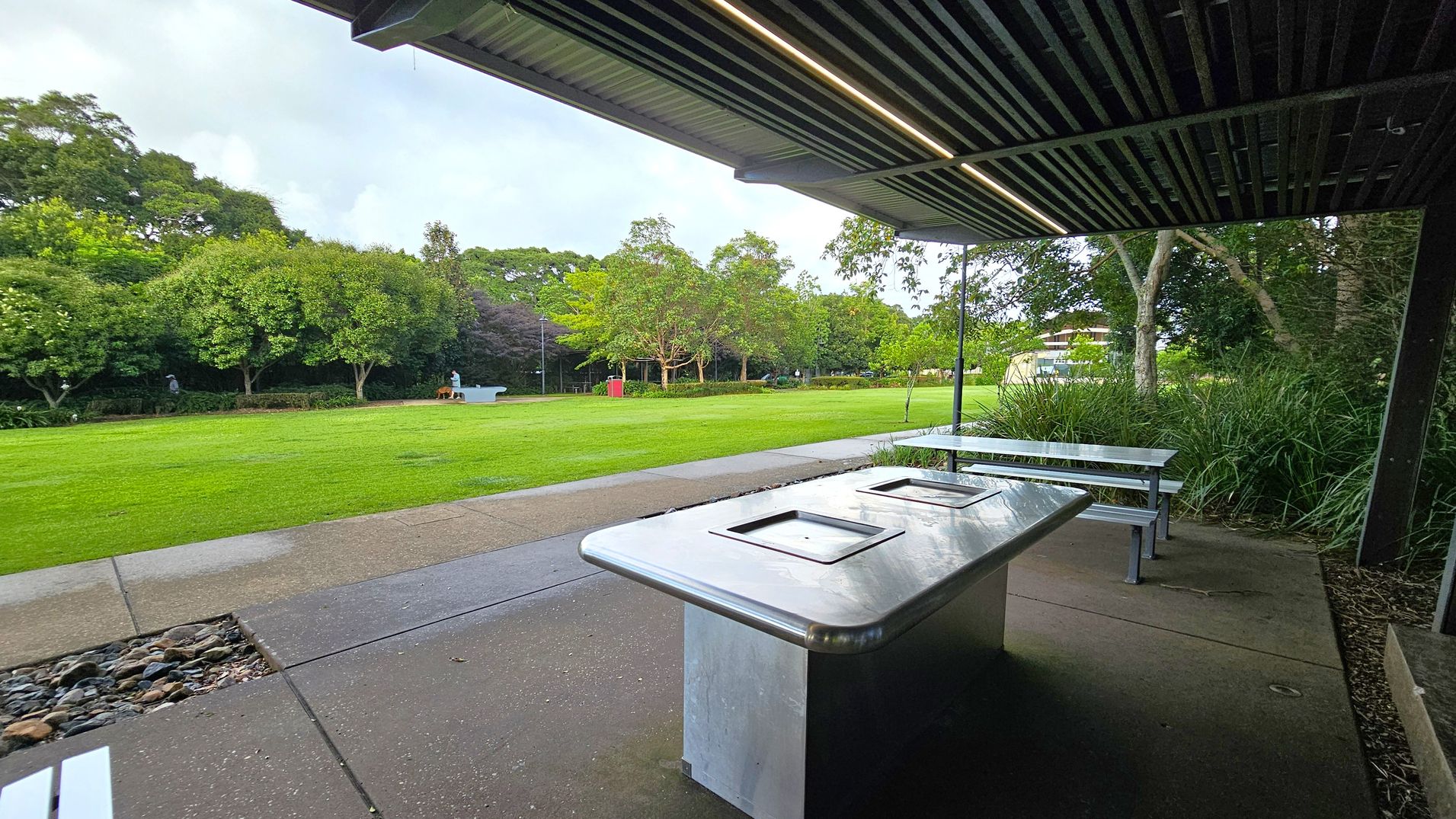 A covered outdoor area with a metal barbecue grill and picnic tables in the foreground. The ceiling is slatted with integrated lighting. The ground is paved with concrete. In the background, there is a large grassy field bordered by trees. A person and a dog are visible near a white bench, and a red bin is situated further back. The scene is surrounded by lush greenery.