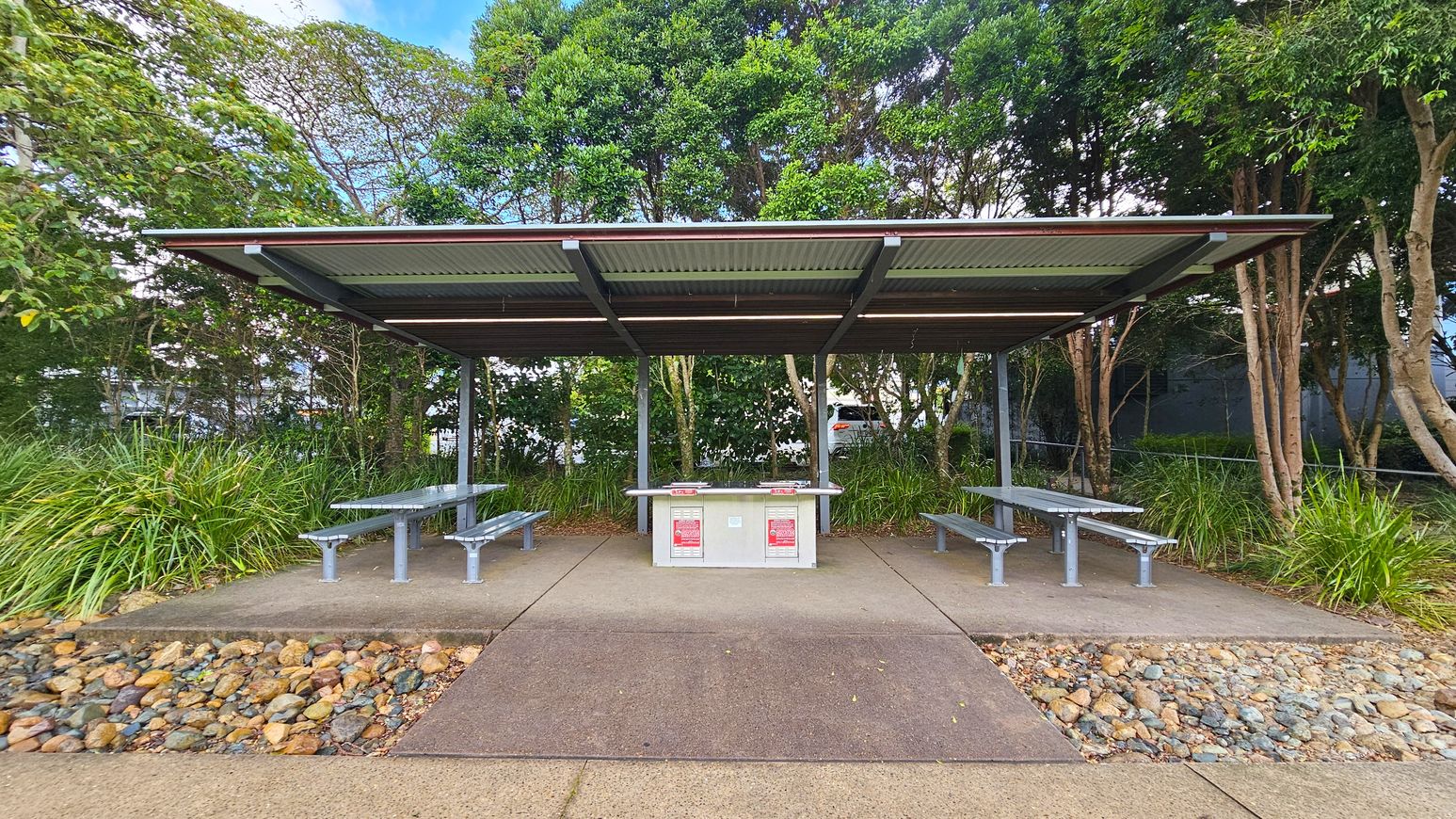 A covered outdoor area with a metal roof and two picnic tables on either side. In the centre, there is a barbecue station with instructions posted on it. The ground is paved with concrete, and there are small rocks lining the edges. In the background, dense green foliage and trees provide a natural setting.