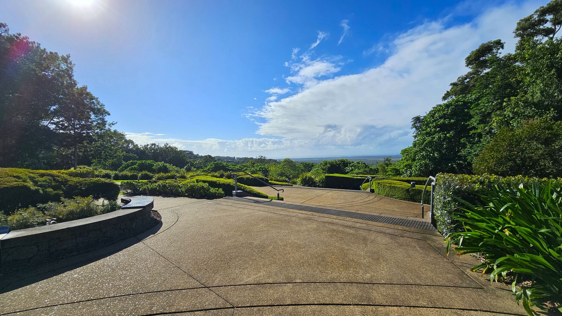 A paved pathway leads into a lush garden area. On the left, there are dense trees and a curved stone bench surrounded by shrubs. On the right, more greenery and manicured bushes line the path. In the centre, the path descends with metal railings on either side. The background features a distant view of the horizon under a partly cloudy sky. The sun is visible in the top left corner, casting light across the scene.