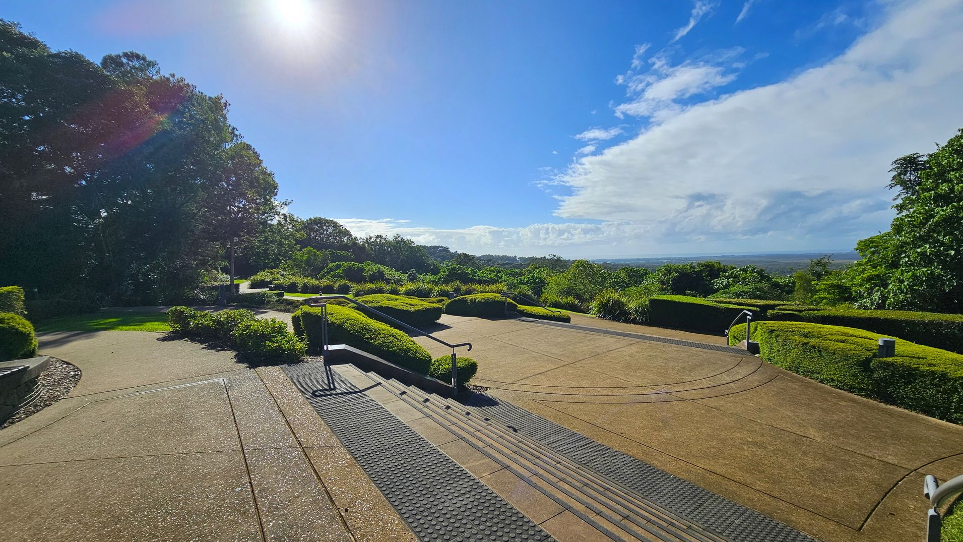 A landscaped garden with a paved pathway and steps in the foreground. The path is bordered by neatly trimmed hedges and lush greenery. On the left, tall trees provide shade, while the right side opens up to a view of distant hills and a clear horizon. The sun is bright in the sky, casting light across the scene. The area is well-maintained, with handrails along the steps and a textured surface for grip.
