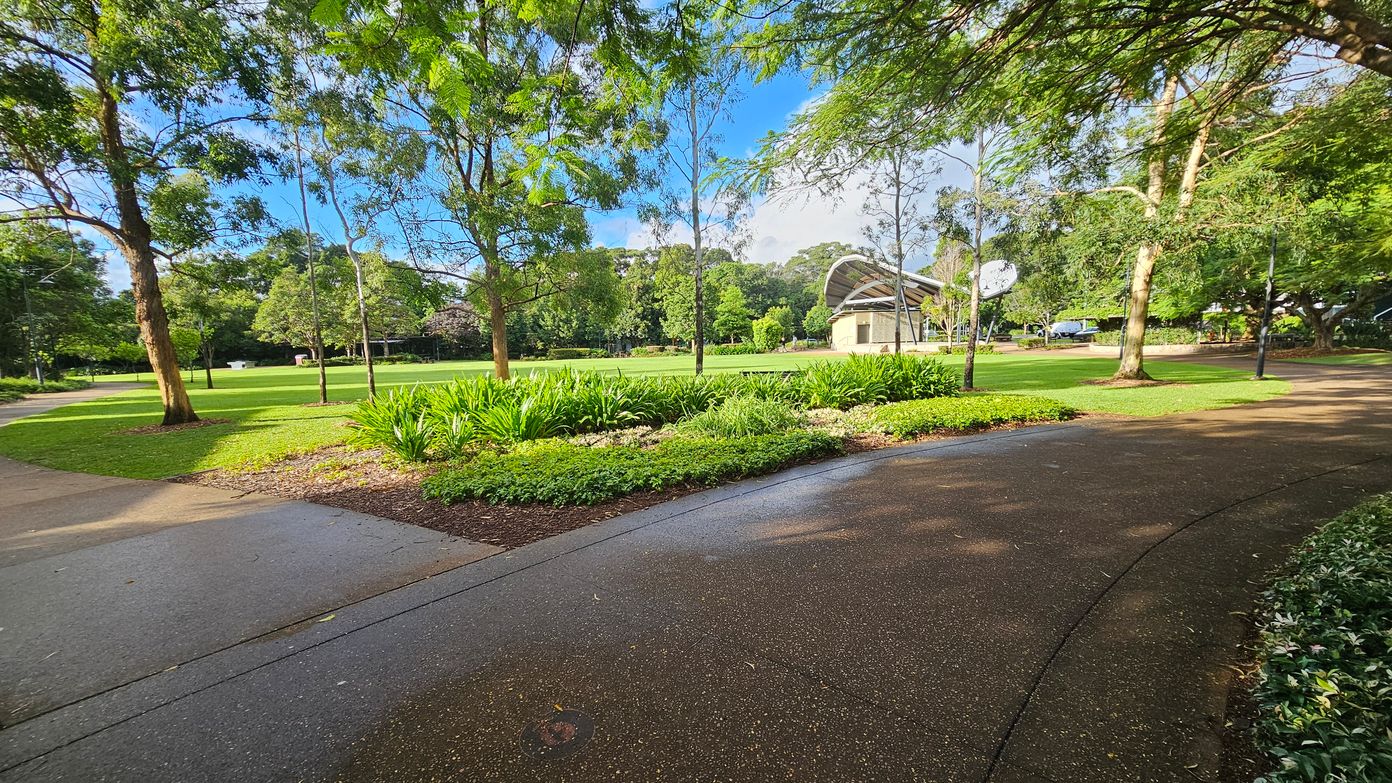 A park scene with a curved pathway in the foreground, bordered by green grass and plants. On the left, tall trees provide shade over the path. In the centre, a grassy area is visible, with more trees in the background. To the right, a structure with a curved roof stands near the path, surrounded by more trees and greenery. The ground is a mix of concrete and brown paving, with some wet patches.