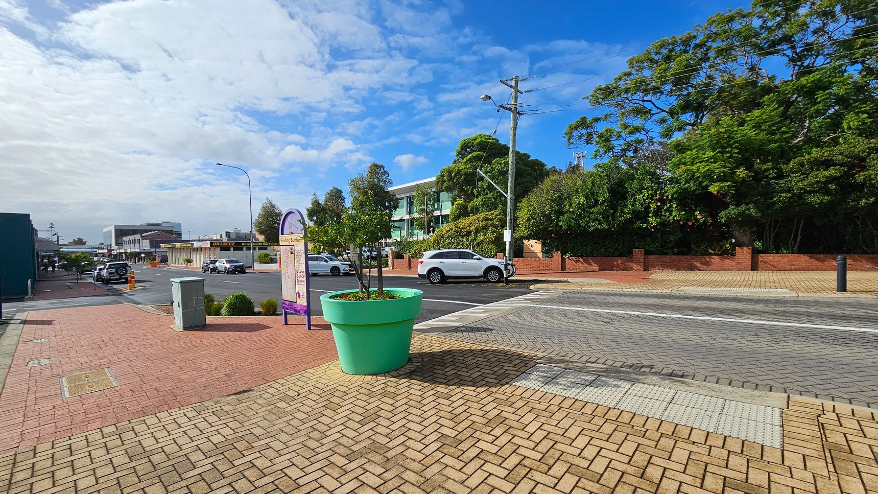 A street scene with a large turquoise planter in the foreground, containing small trees. The pavement is made of light brown bricks. To the left, a sign with a map stands near a grey utility box. In the background, a white car is parked on the road, with a building partially obscured by trees. The right side features a brick wall and dense greenery. The sky is partly cloudy.