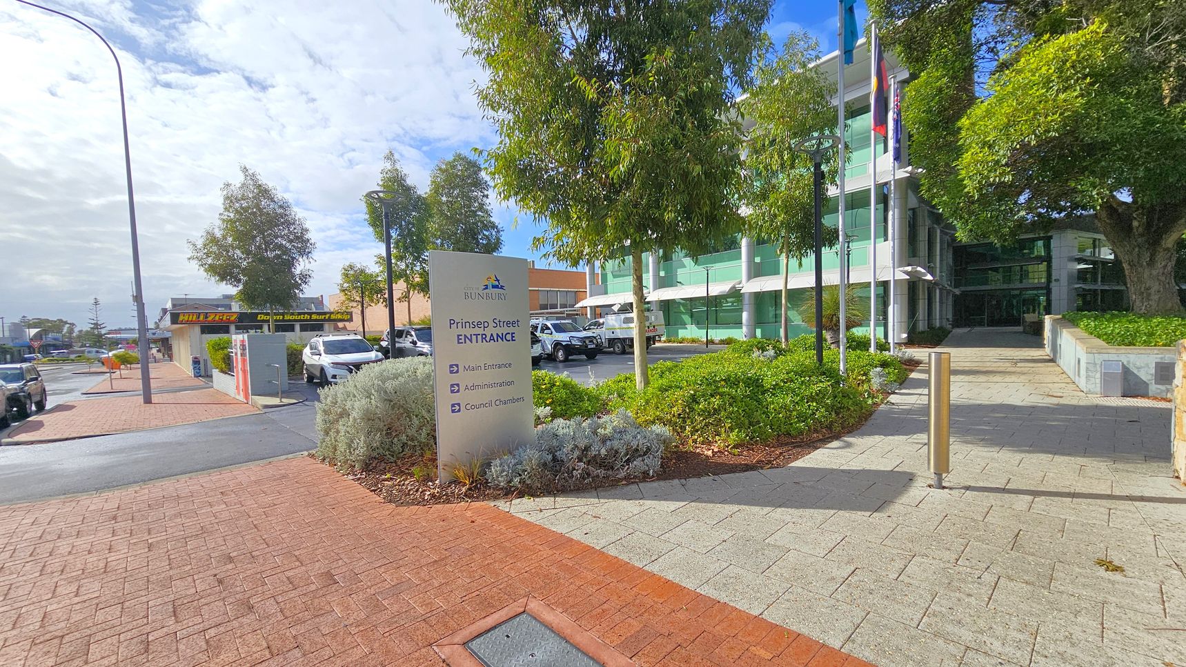 A paved walkway leads to a modern glass building on the right, partially obscured by trees and greenery. A sign in the foreground reads "Prinsep Street Entrance" with directions. On the left, a street with parked cars and a shop sign is visible. The pavement is a mix of red and grey bricks, and there are tall street lamps along the path. Flags are flying near the building entrance.