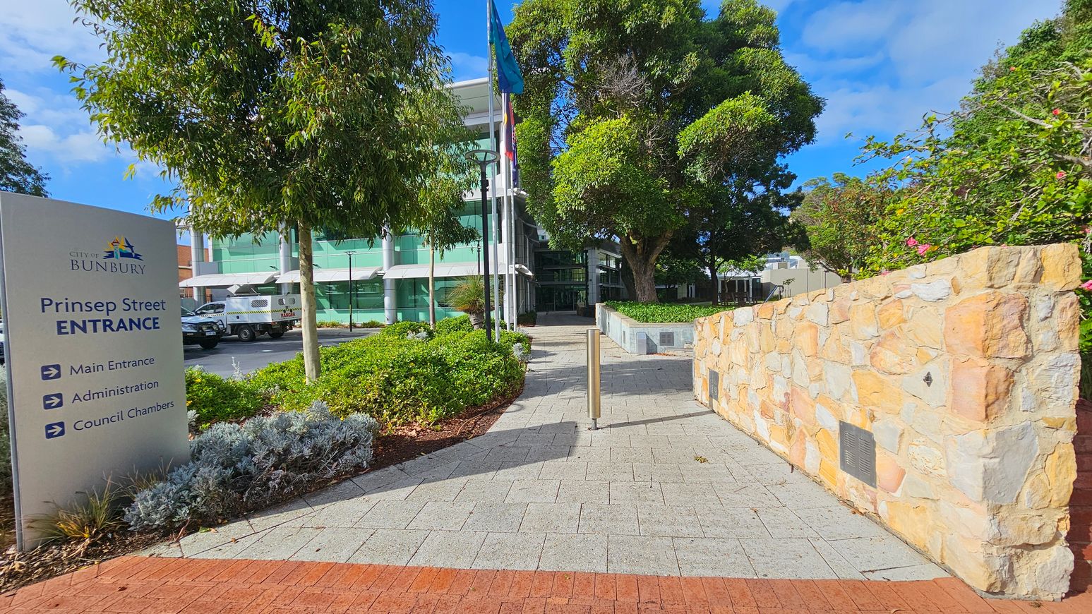 A paved walkway leads to a modern building with green-tinted glass windows in the background. On the left, a sign reads "City of Bunbury, Prinsep Street Entrance" with directions. A tree and a parked vehicle are nearby. On the right, a stone wall with plaques runs alongside the path, bordered by green shrubs and flowering plants. Several flags are visible near the building entrance. The area is well-landscaped with trees and bushes.