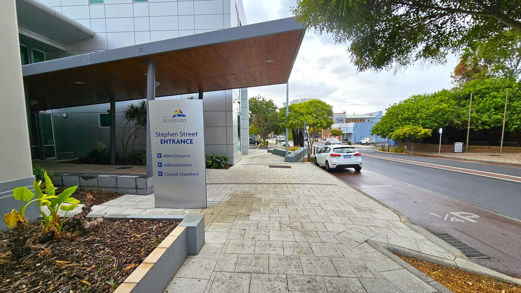A paved walkway leads to a building with a sign reading "City of Bunbury, Stephen Street Entrance" on the left. The sign directs to the main entrance, administration, and council chambers. A modern building with a slanted roof is adjacent. On the right, a street with a parked white car and lush green trees lines the road. The pavement is light grey with a subtle pattern, and the area is well-maintained with small plants in a bed near the sign.