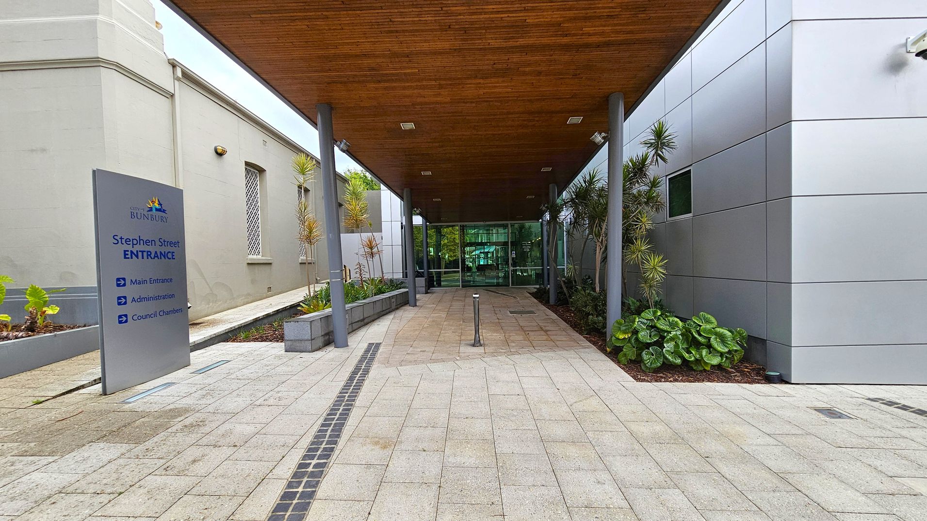 A covered walkway leads to a glass entrance. On the left, a grey building with barred windows and a sign reading "Stephen Street Entrance" with directions to the main entrance, administration, and council chambers. On the right, a modern building with grey panels and plants along the side. The ceiling is wooden, and the floor is paved with light-coloured tiles.