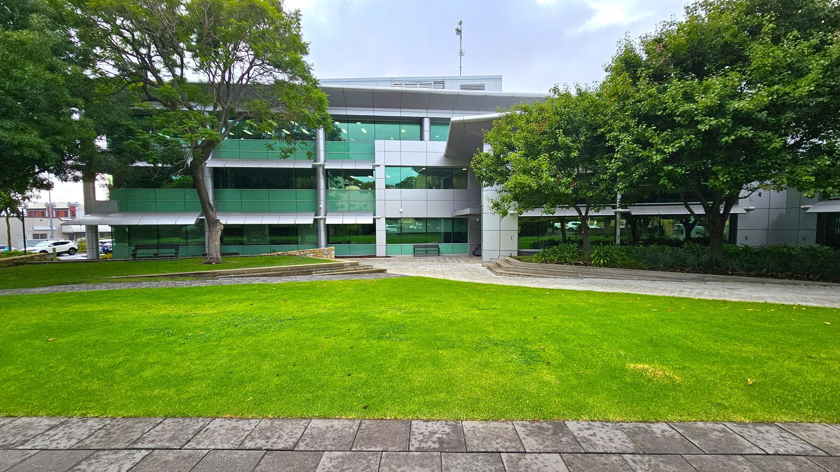 A modern building with large green-tinted windows and a grey facade is in the background. In the foreground, there is a well-maintained lawn bordered by a paved walkway. Trees with dense foliage are on both sides, providing shade. Benches are placed near the building, and there are landscaped areas with plants. The scene is calm and organised, with a mix of natural and architectural elements.