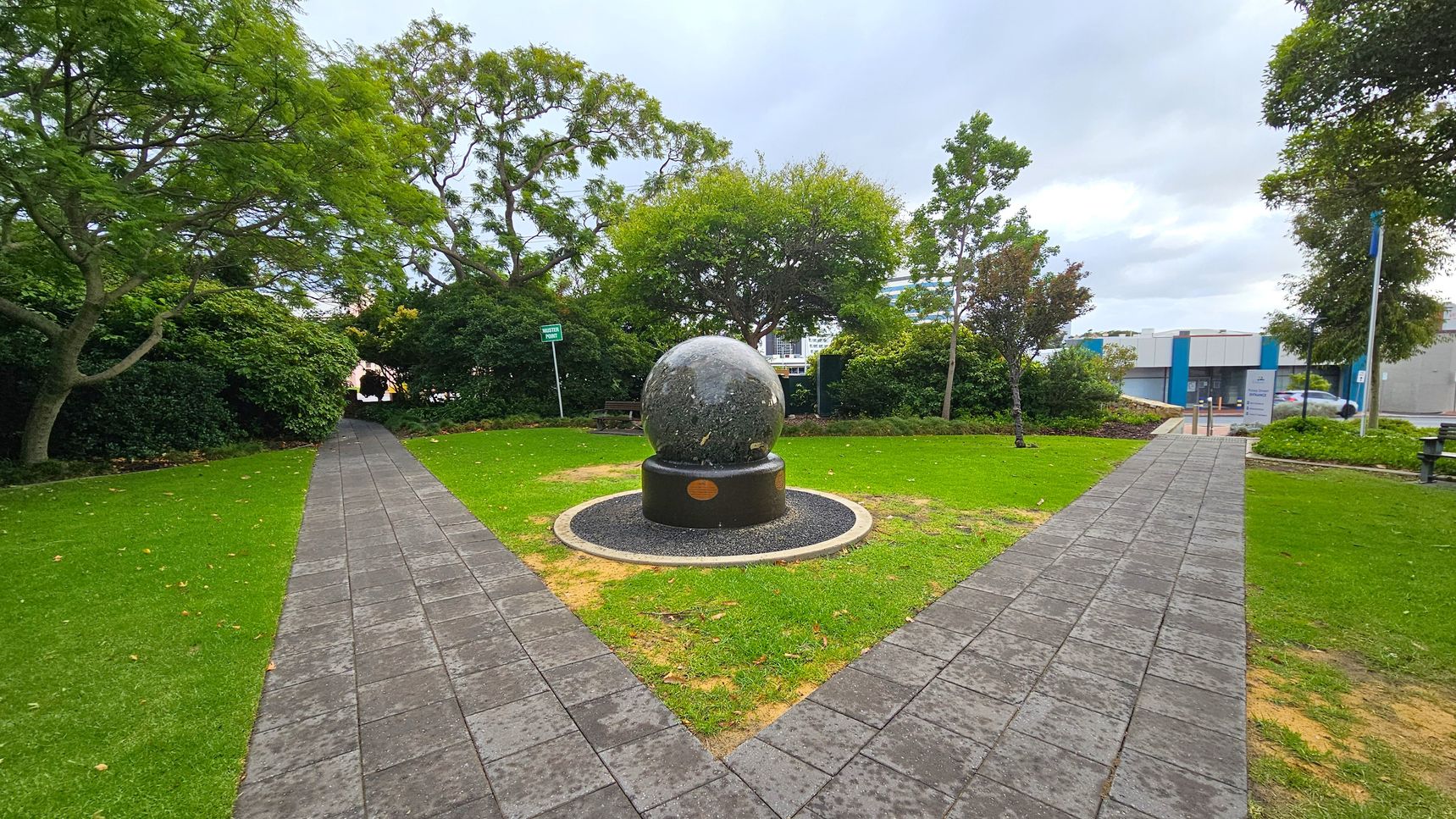 A spherical stone sculpture is centred on a circular base in a small park. Two paved paths intersect at the sculpture, forming a V-shape. Lush green grass surrounds the paths, with trees and bushes lining the edges. In the background, a modern building with blue accents is visible. A bench is partially visible to the left, and a signpost is near the centre. The sky is overcast.