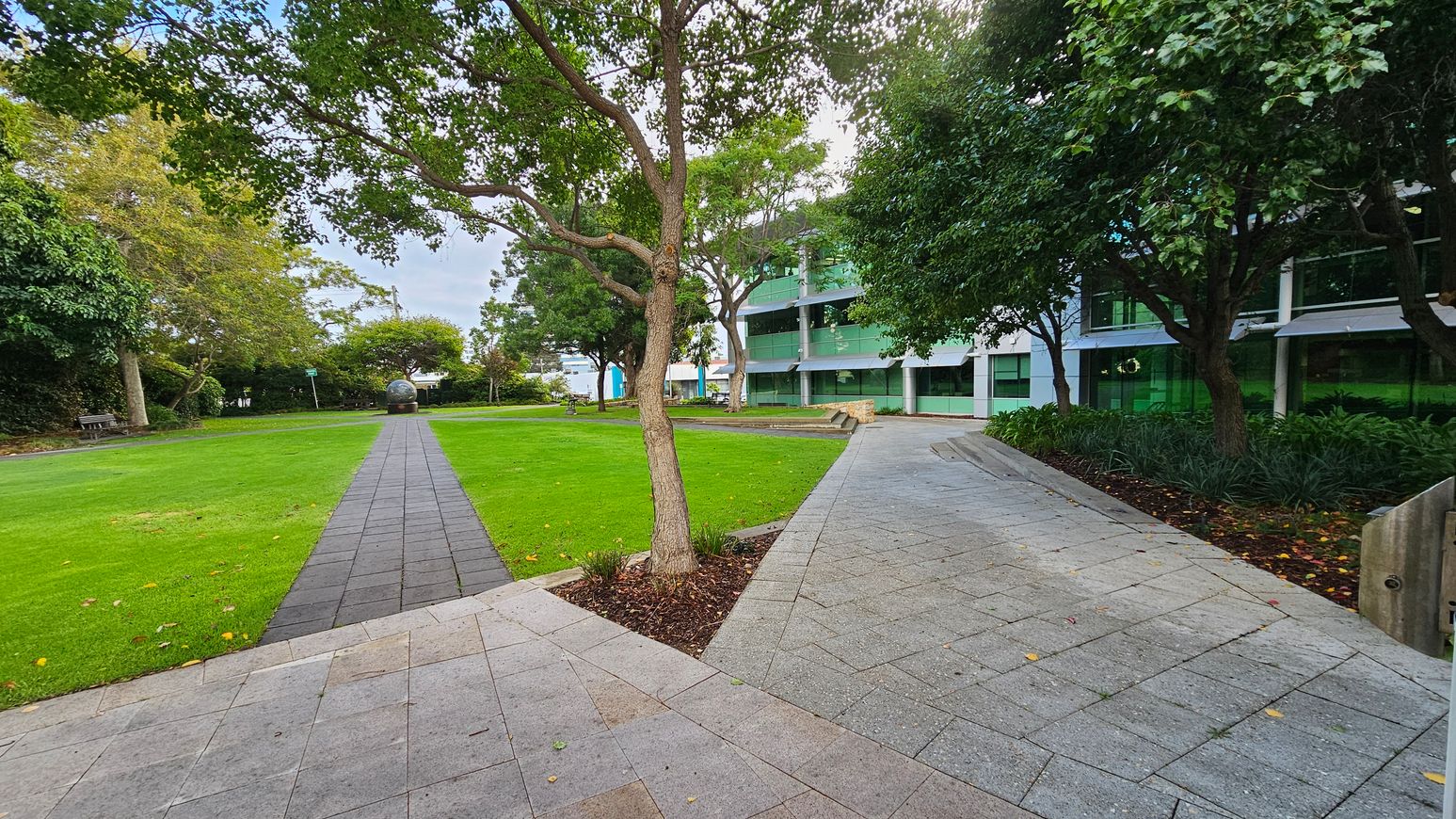 A paved walkway runs through a grassy area with trees on both sides. On the left, a bench is partially visible under the trees. In the centre, a spherical sculpture is situated at the end of the path. On the right, a modern building with large green-tinted windows is partially obscured by trees and shrubs. The ground is covered with grey paving stones, and the area is surrounded by lush greenery.
