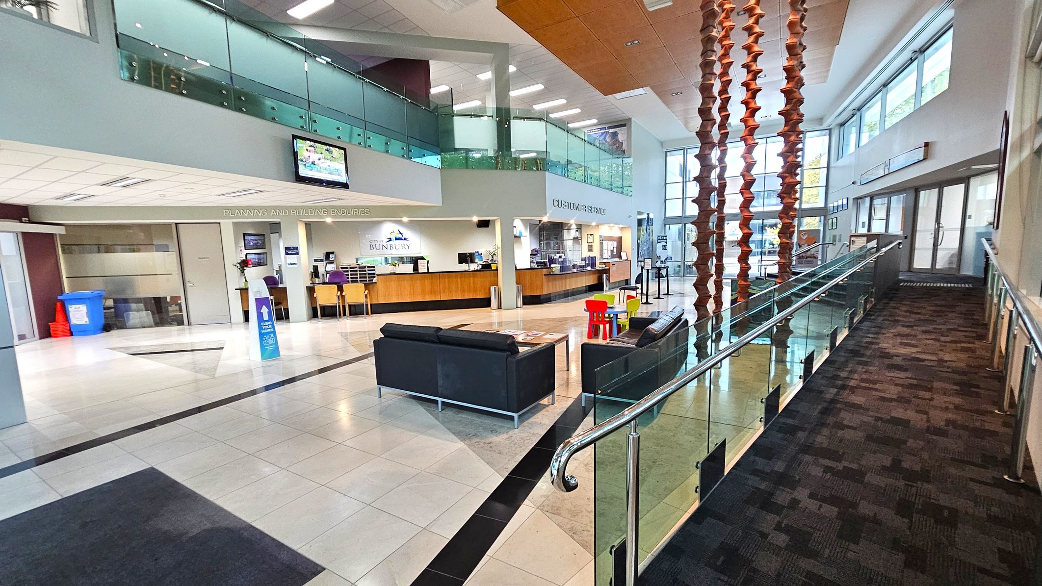 A spacious interior with a modern design. On the left, there is a reception area with a counter and chairs, labelled "Planning and Building Enquiries". Above, a glass-panelled balcony overlooks the space. In the centre, black sofas and a small table with colourful children's chairs are arranged on a tiled floor. On the right, a carpeted ramp with glass railings leads to a set of glass doors. Large windows on the right wall allow natural light to fill the area.
