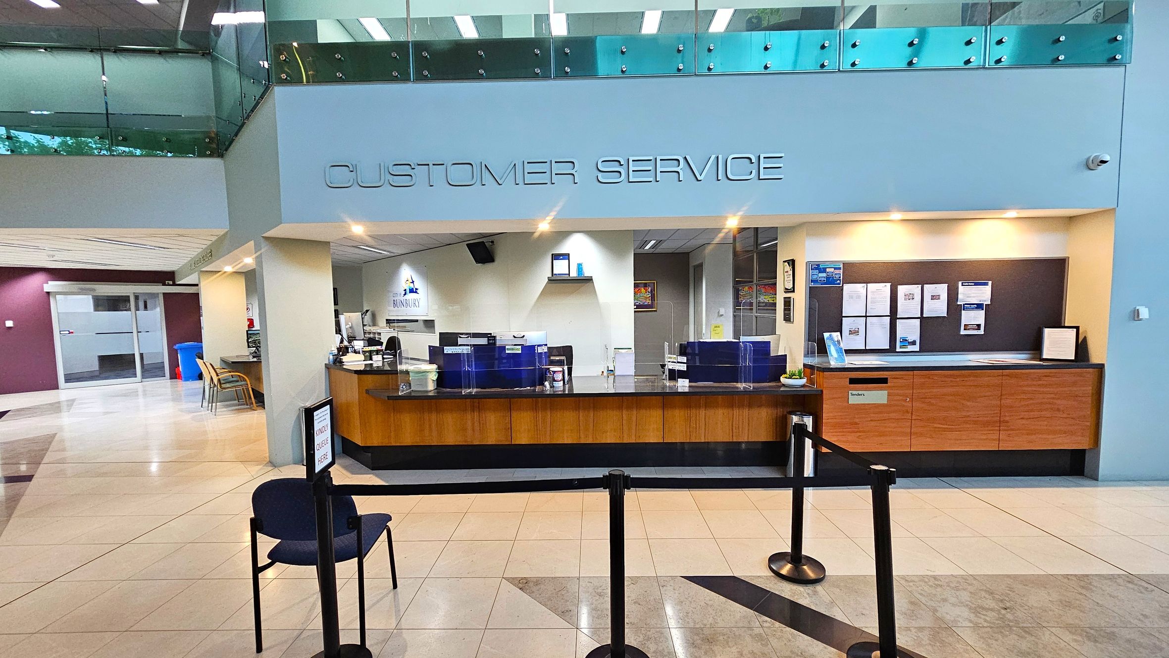 A customer service area with a wooden counter and clear partitions. Above, "CUSTOMER SERVICE" is written on the wall. Behind the counter, there are computers and a sign with "BUNBURY" visible. On the right, a notice board displays various papers. The floor is tiled, and a single chair is placed in front of a queue barrier. The ceiling has recessed lighting, and a glass railing is visible on the upper level.