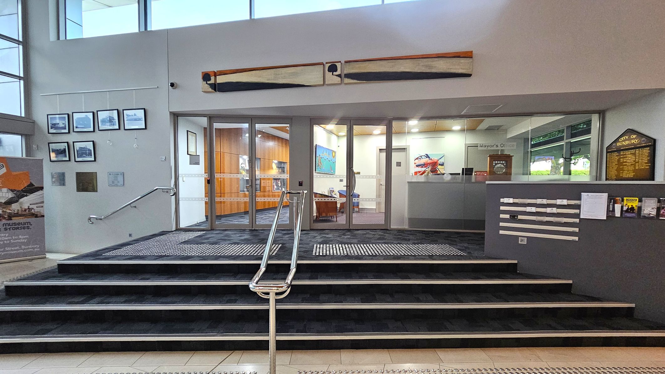 A modern interior with a set of stairs leading up to a glass entrance. On the left, framed photographs are displayed on the wall above a handrail. A sign with text is positioned on the floor. Above the entrance, a long artwork is mounted. To the right, a grey wall features a plaque and several brochures. Inside, the space is well-lit with wooden panelling and colourful artwork visible. A sign indicates the "Mayor's Office". The floor is tiled with a textured mat at the entrance.