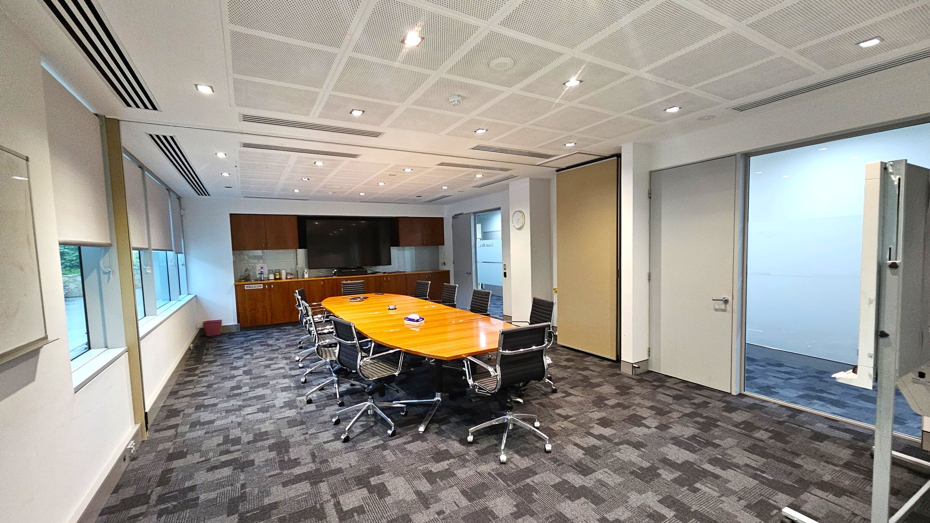 A modern conference room with a long wooden table in the centre, surrounded by black office chairs on wheels. On the left, large windows with blinds allow natural light in. The back wall features a large screen and a wooden cabinet with various items. The right side has a door leading to another room and a whiteboard. The floor is carpeted with a grey and purple pattern, and the ceiling is white with recessed lighting.