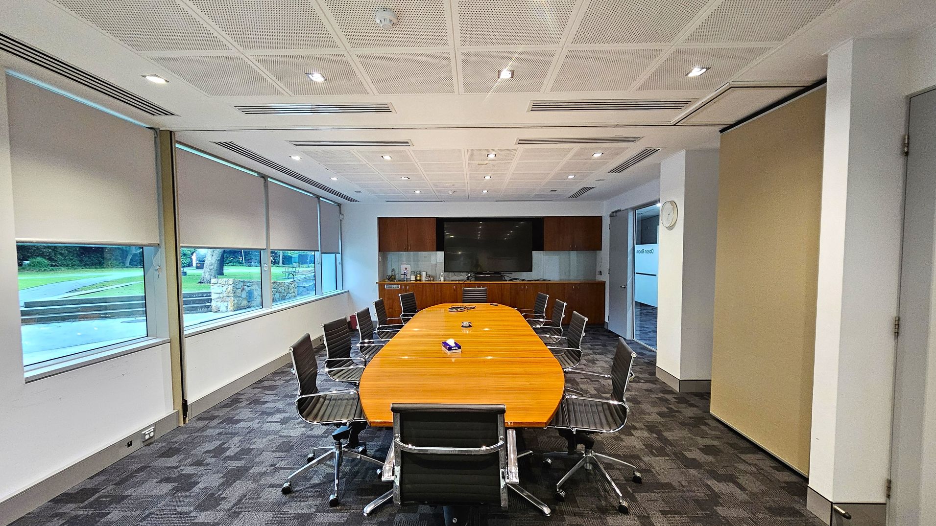 A conference room with a long, oval wooden table surrounded by black office chairs on a patterned carpet floor. The left side features large windows with roller blinds partially down, revealing a view of a grassy outdoor area. The back wall has a large flat-screen TV mounted above a wooden cabinet. The ceiling is white with recessed lighting. On the right, a wall clock is visible above a doorway.