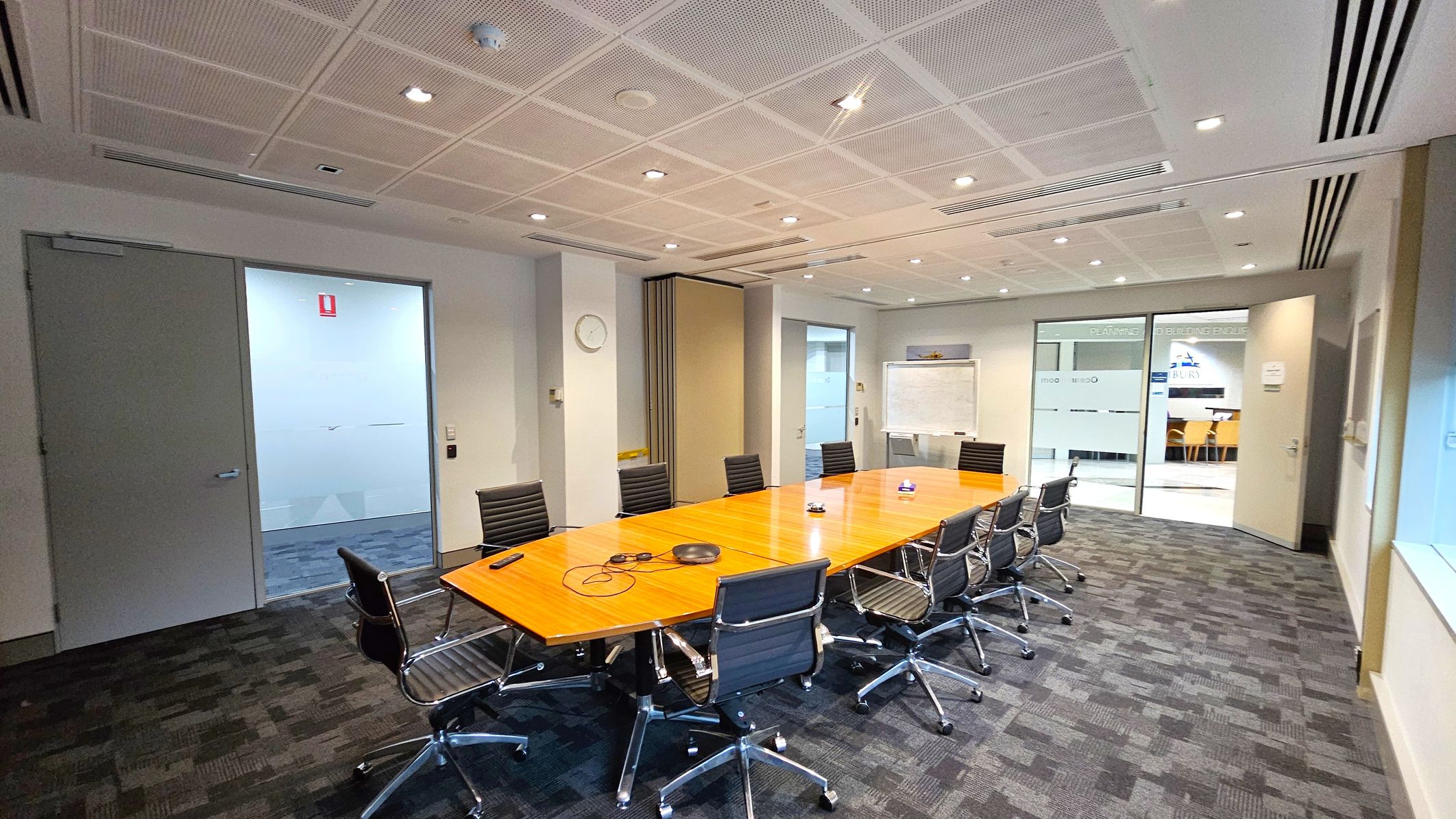 A conference room with a long wooden table surrounded by black office chairs on a dark carpeted floor. On the left, there is a closed door and a wall clock. In the centre, a whiteboard is visible against the wall. On the right, glass doors lead to another area with visible signage and a glimpse of a seating area. The ceiling is fitted with recessed lighting and air vents.