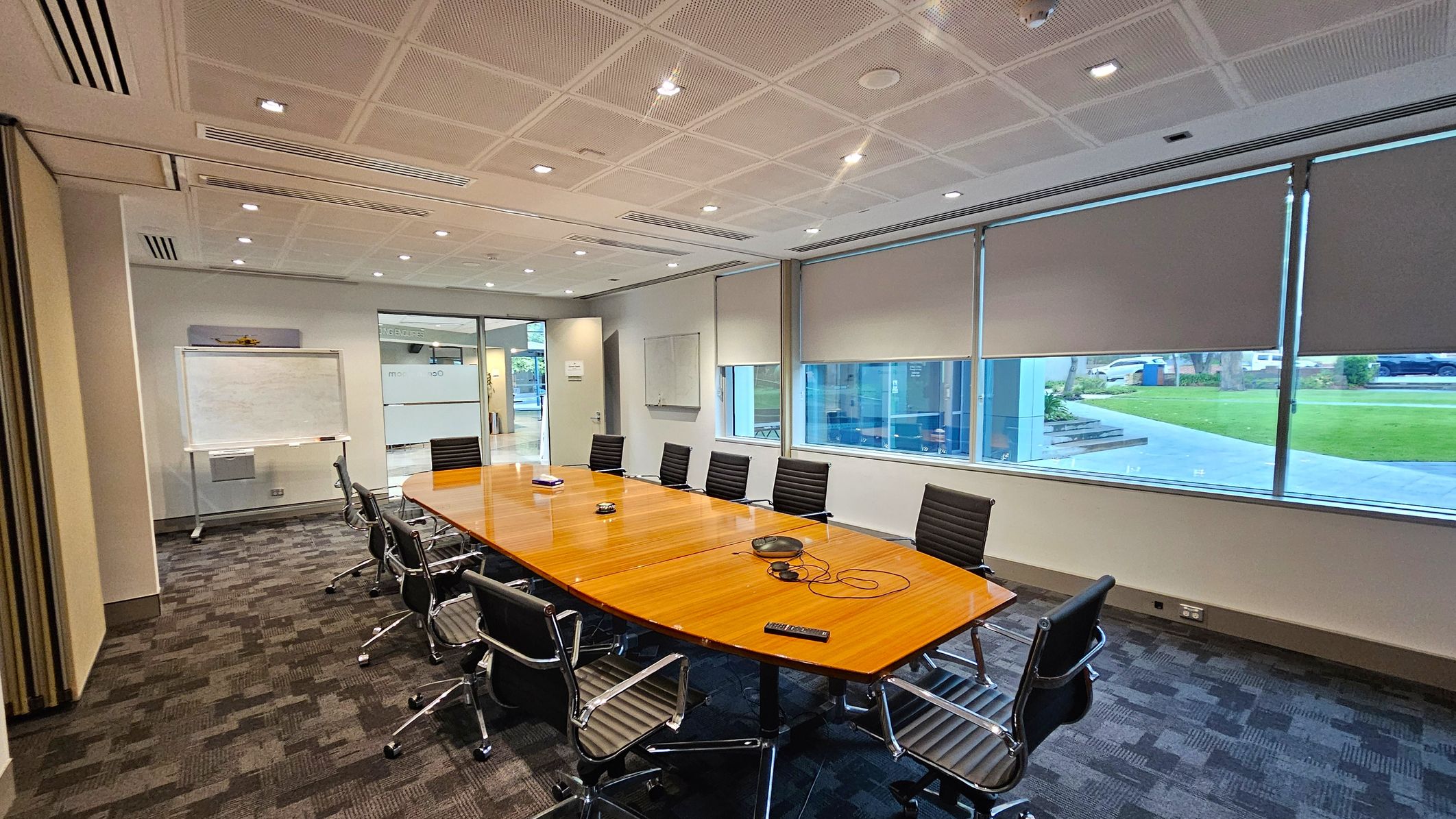 A modern conference room with a large wooden table in the centre surrounded by black office chairs on wheels. On the left, there's a whiteboard on a stand. The right side features large windows with partially drawn blinds, offering a view of a grassy area outside. The floor is carpeted with a grey and black pattern, and the ceiling is fitted with recessed lighting and air vents.