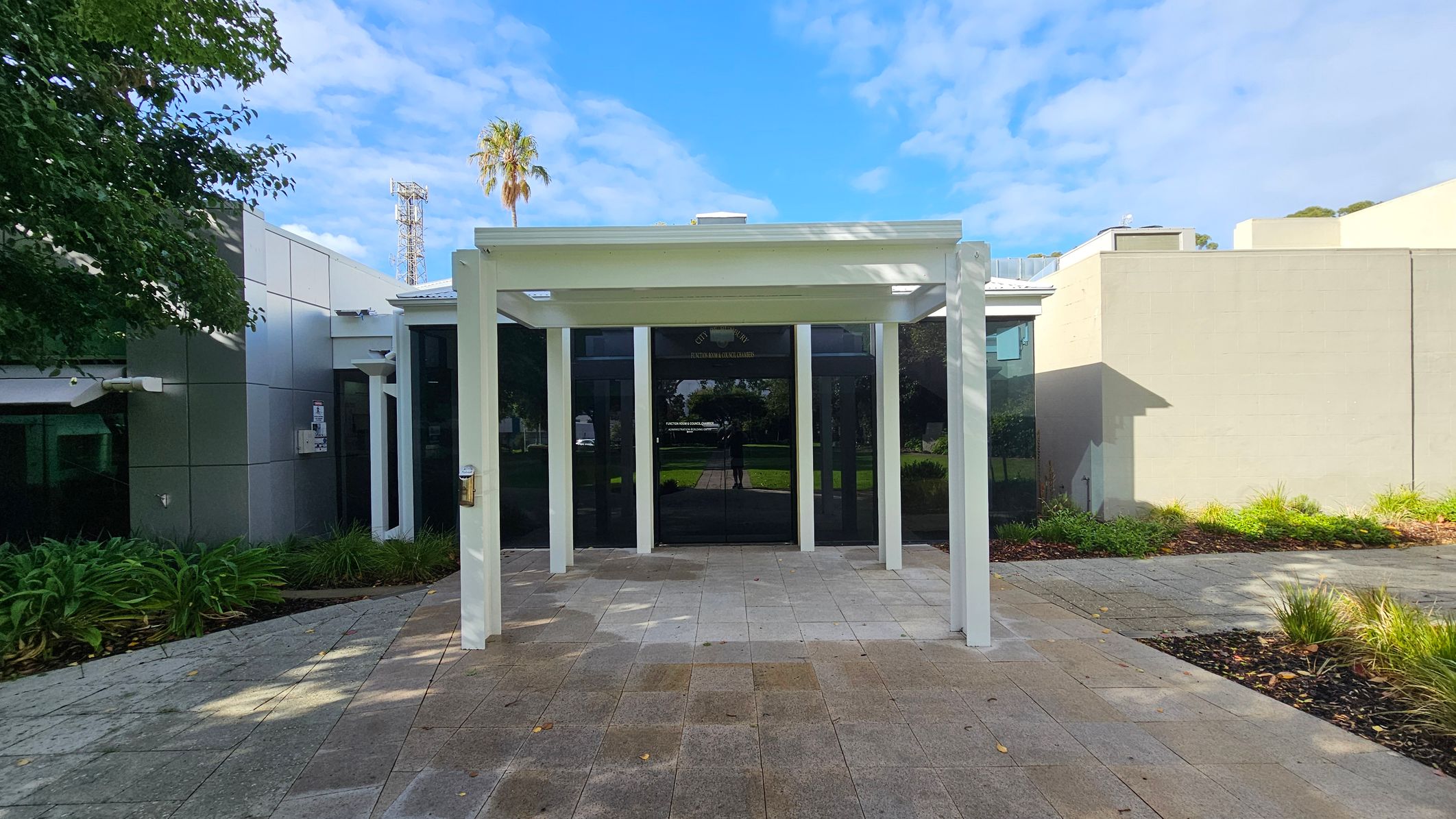 A modern building entrance with a white pergola structure in the foreground. The ground is paved with grey and brown tiles. On the left, there are green plants and a tree partially visible. The building has a glass facade with reflections and a light grey wall on the right. In the background, a palm tree and a tall metal structure are visible against a blue sky.