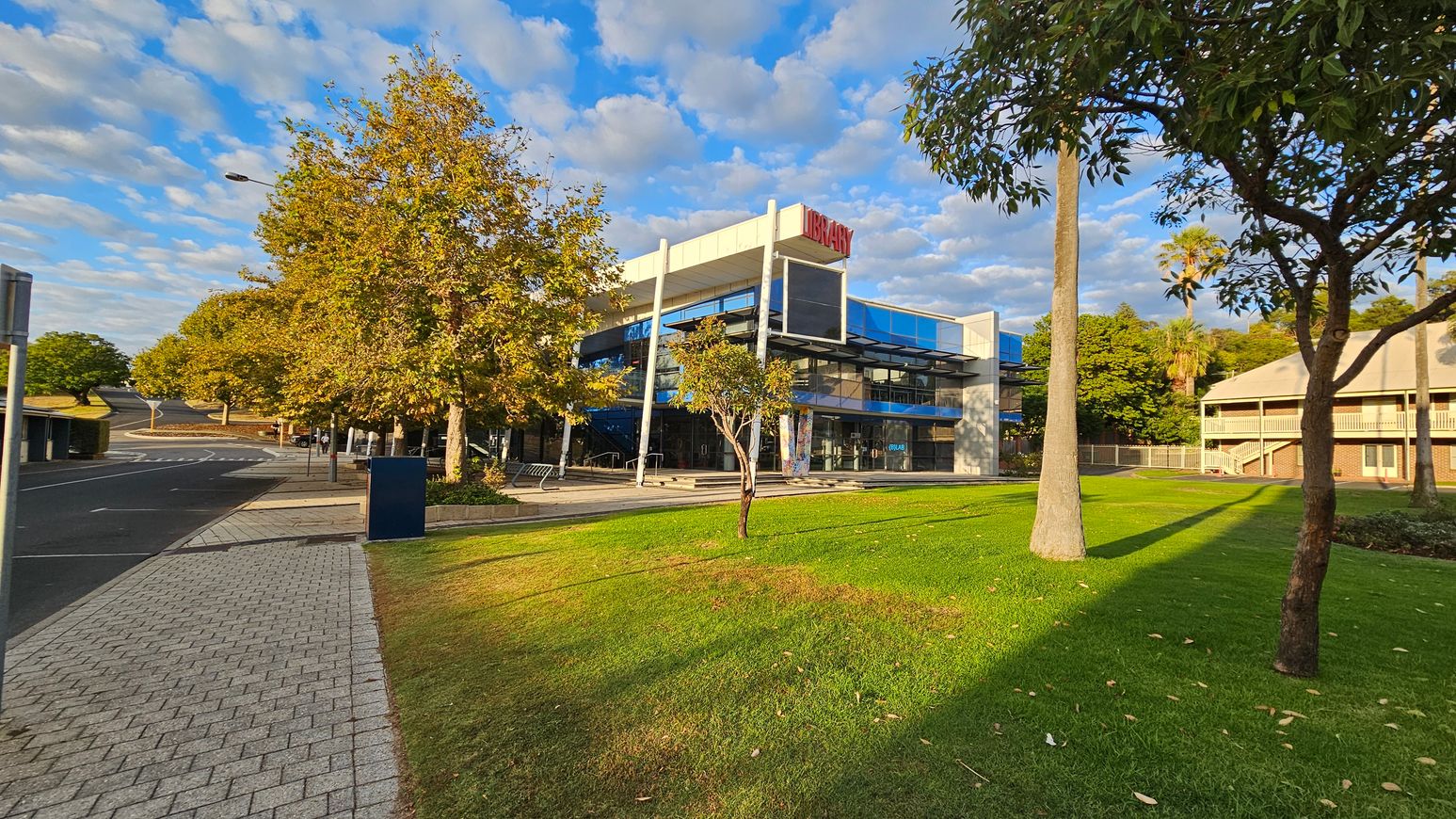 A modern library building with a sign on top is situated in the centre, featuring large glass windows and a white facade. To the left, a paved walkway runs alongside a road lined with trees showing autumn foliage. On the right, a grassy area with scattered trees leads to another building with a sloped roof and balconies. The sky is partly cloudy, casting shadows on the ground.